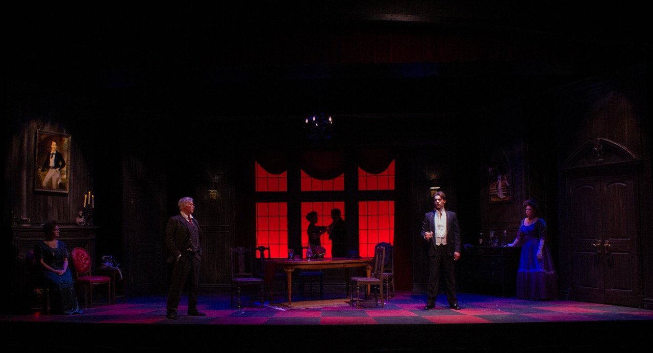 Scene from a theatrical play set in a dark room with five people; two of them appear as silhouettes behind a window with red lighting, and three are in the foreground, dressed in vintage attire, standing on a stage with dark wooden walls and old-fash
