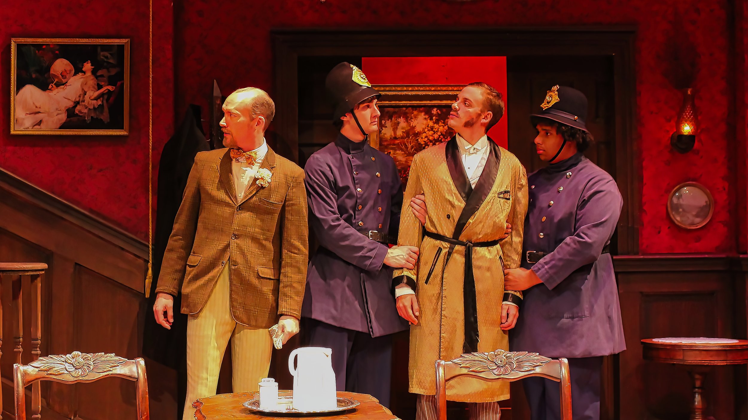 Four actors dressed in period costumes standing in a room with red walls and vintage decor, appearing to be in a theatrical performance or play.