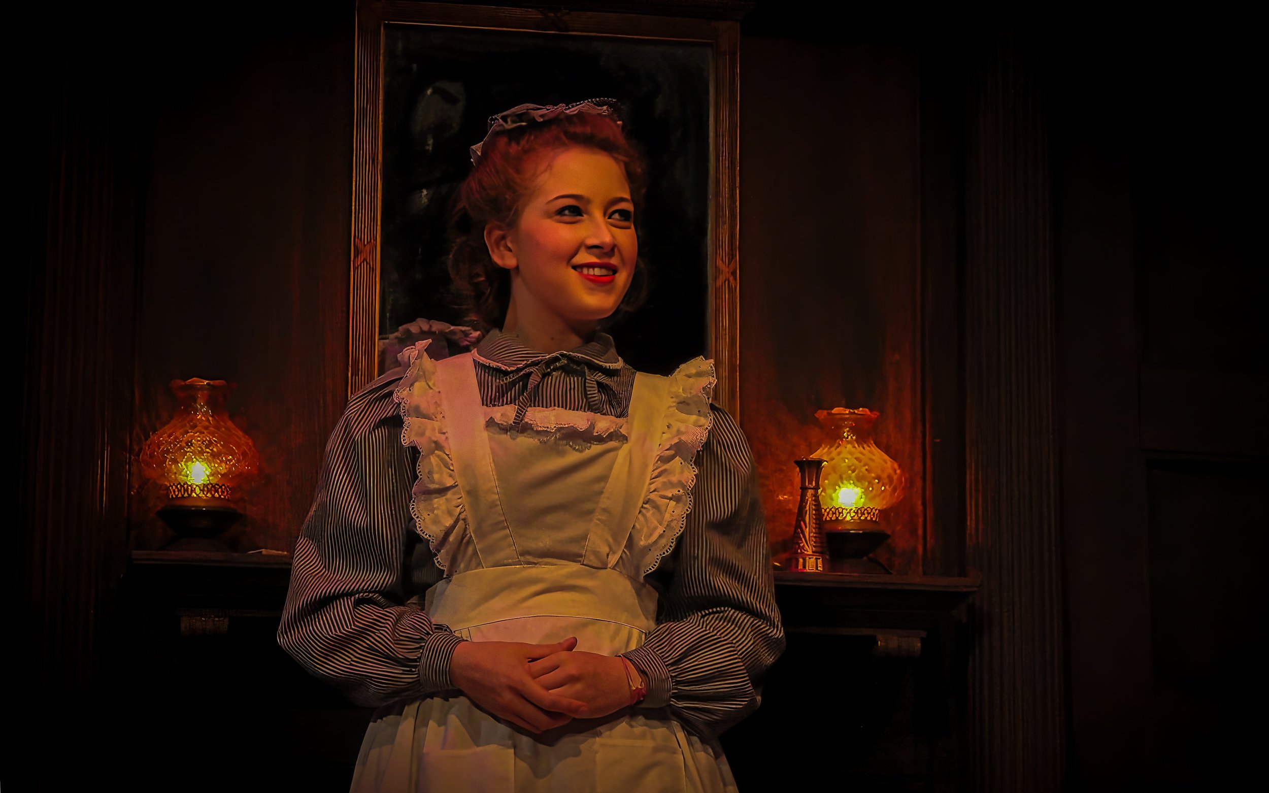Young woman dressed as a maid with a striped blouse and white apron, standing in front of a dark wooden background with a mirror, two glowing lamps, and a small decorative Eiffel Tower figurine.