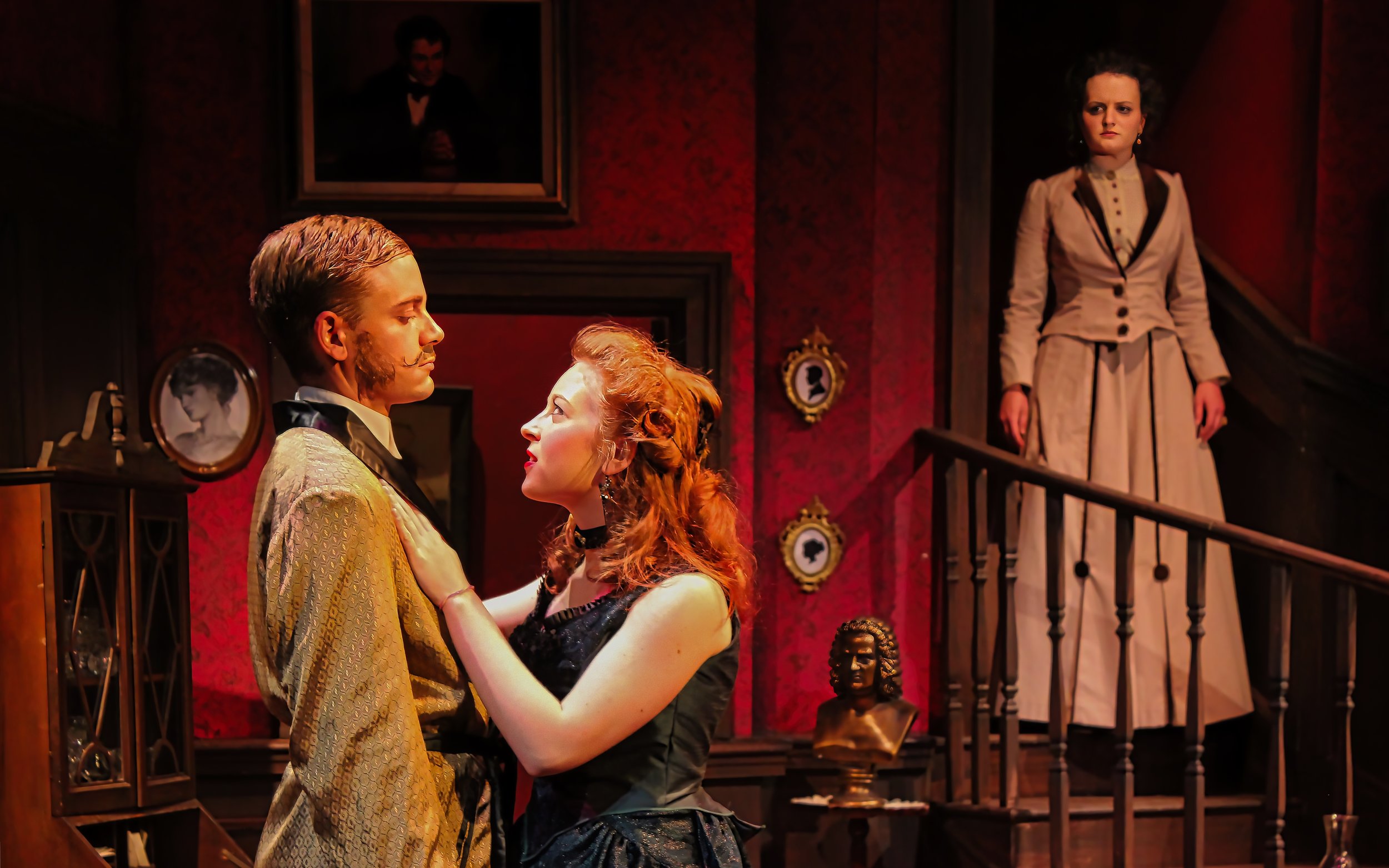 A man and woman dance closely in a vintage-themed room, with an older woman standing on a staircase observing them. The room features red wallpaper, framed portraits, and a bust on a pedestal.