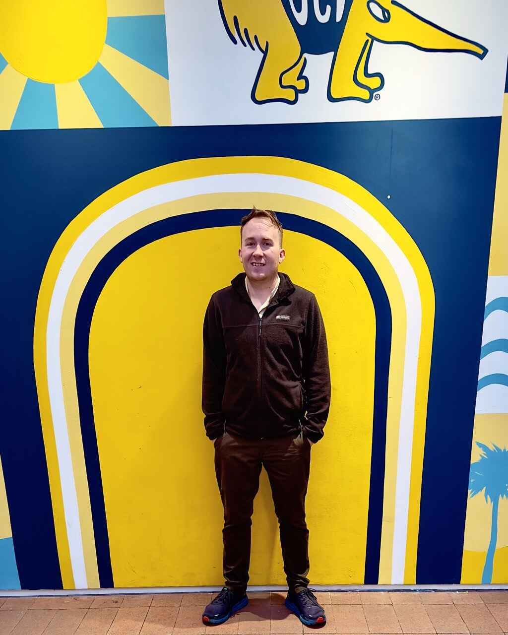Person standing in front of a colorful mural with blue, yellow, and white design and palm tree elements.