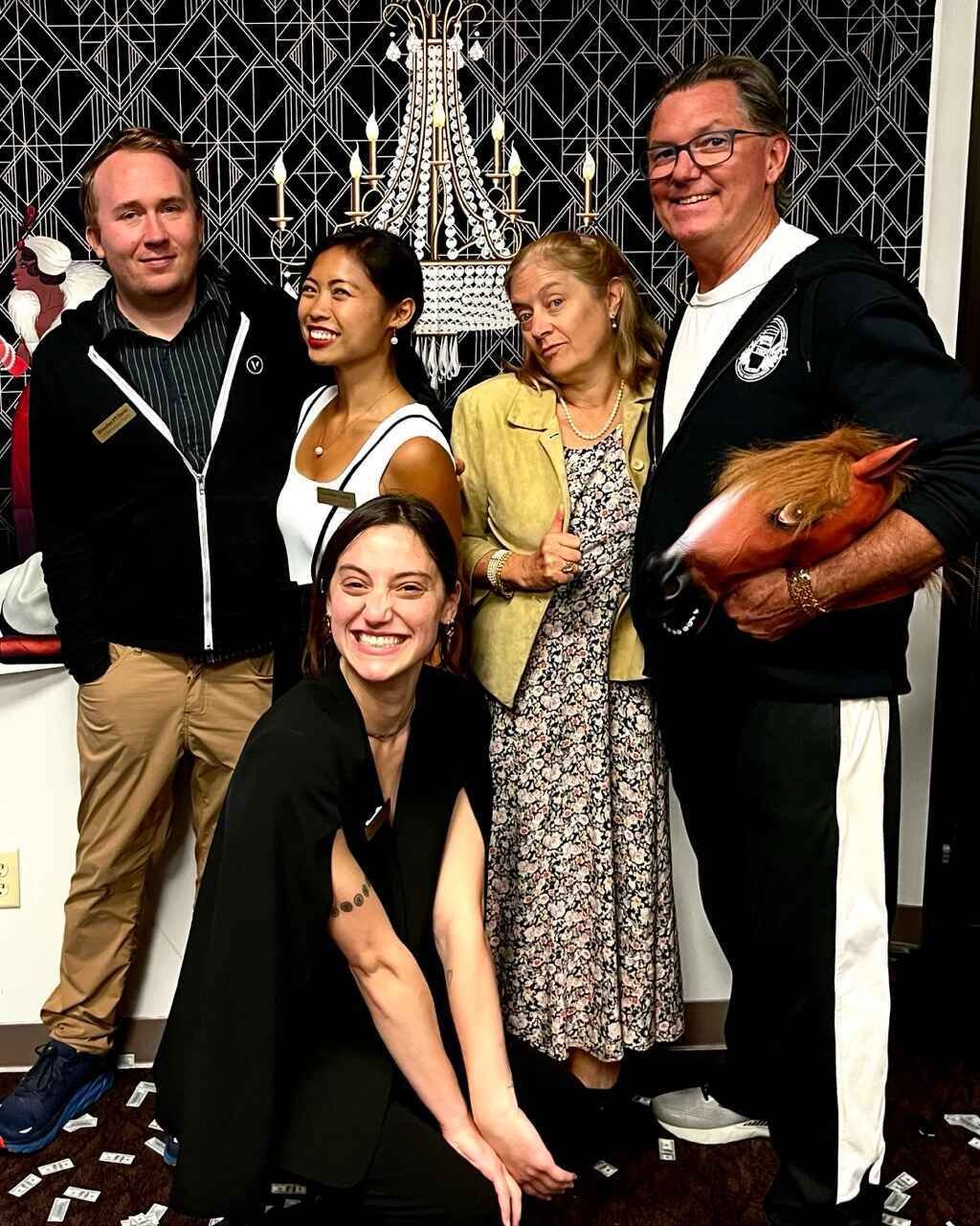 Group of five people smiling and posing indoors, with one person holding a horse mask. They are standing in front of a decorative black and white backdrop with a chandelier design. Confetti is scattered on the floor.