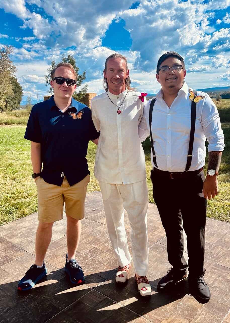 Three people posing together outdoors on a sunny day, with one person wearing a white outfit and the others in casual attire, standing on a tiled area with grass and a blue sky in the background.