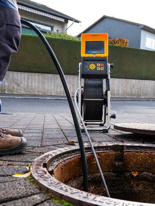A person using a sewer camera inspection device to examine a sewer manhole on a sidewalk outside residential houses.