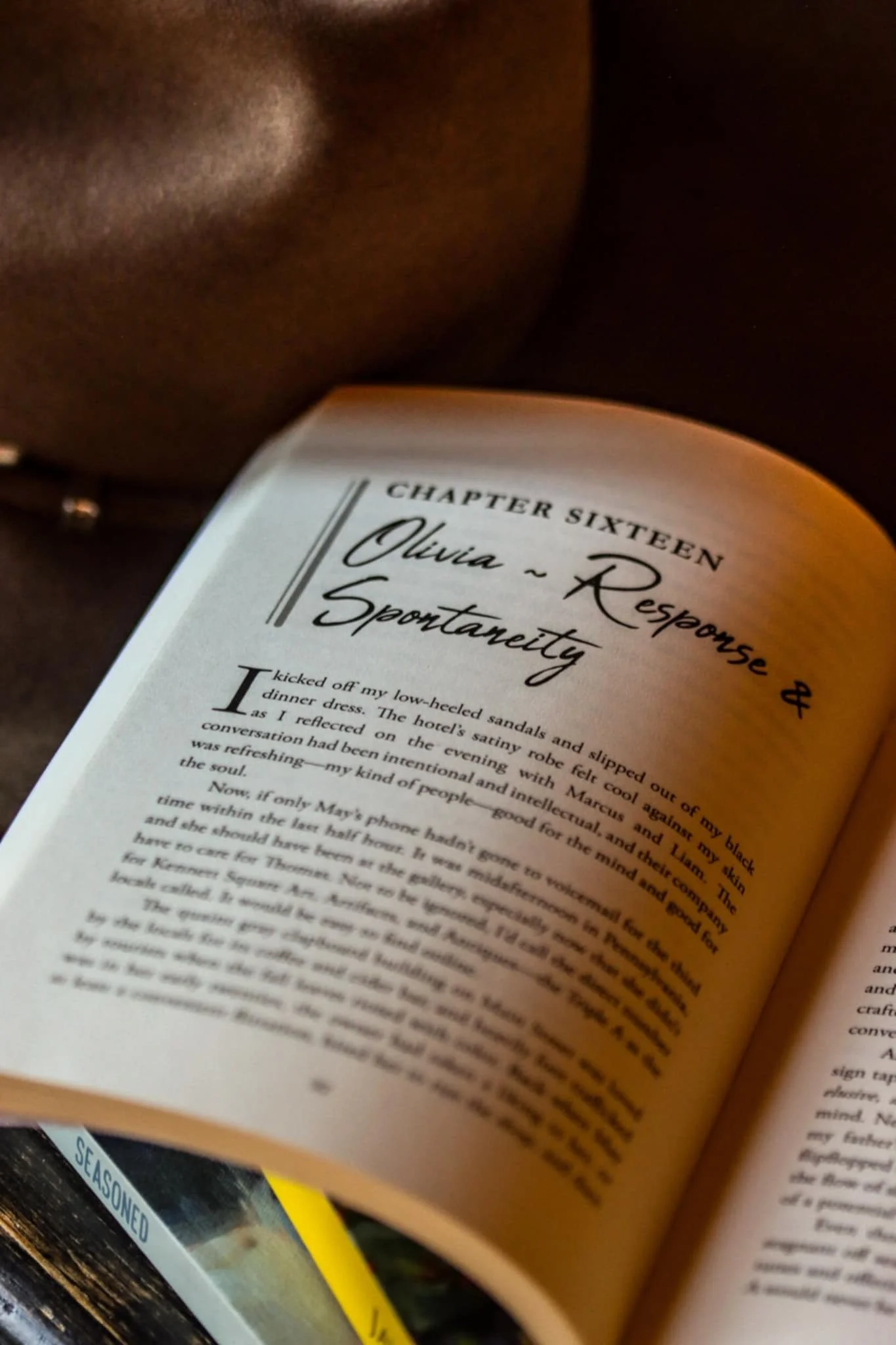 A close-up of a book open to a chapter titled 'Chapter Sixteen' with the header 'Olivia - Response & Spontaneity.' The page features printed text, and the person's face is partially visible in the background, showing dark skin and wearing a necklace.
