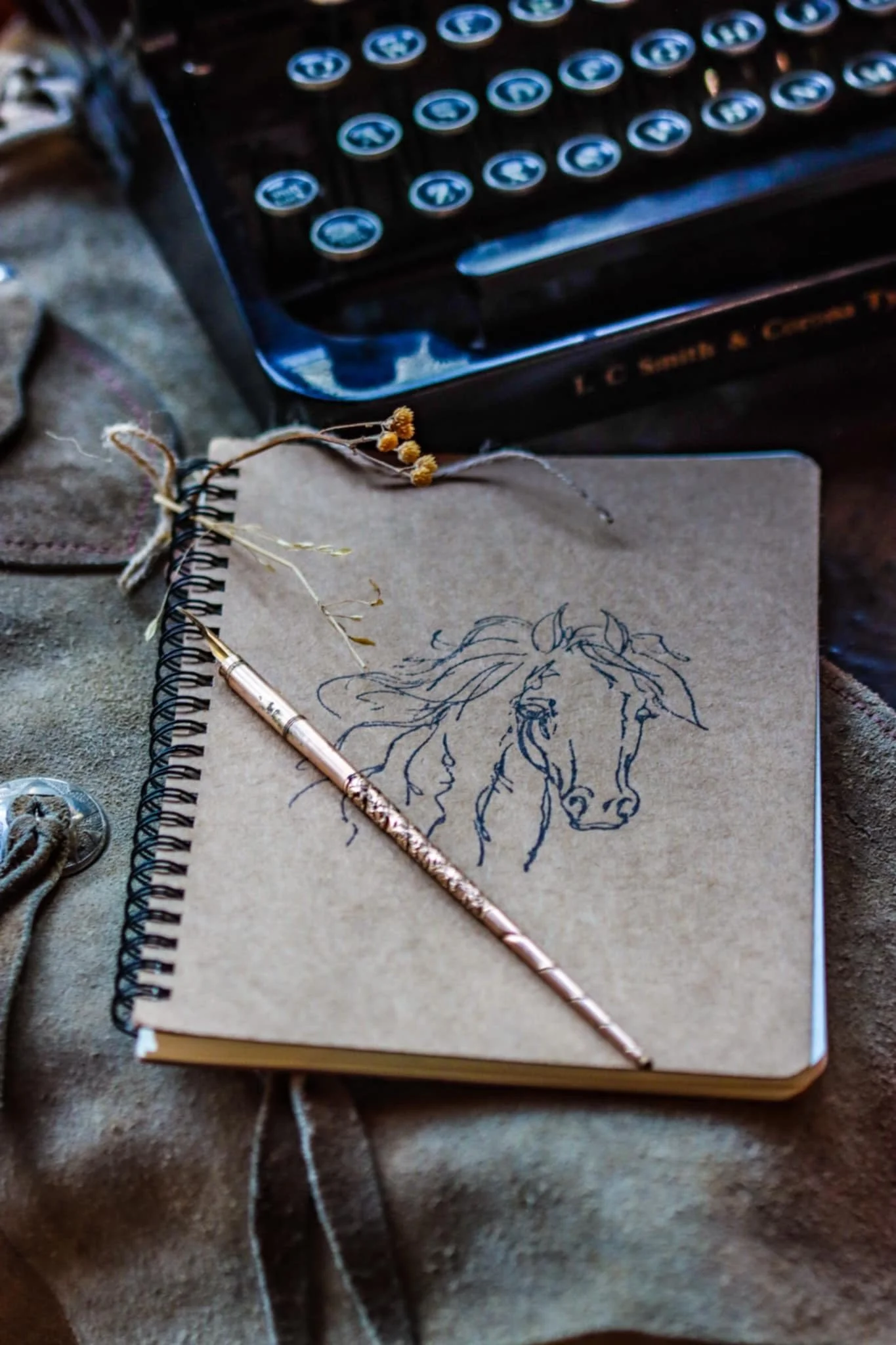 A spiral notebook with a drawing of a horse and a woman's face on brown paper, a decorative pen, dried flowers, and a vintage typewriter in the background.