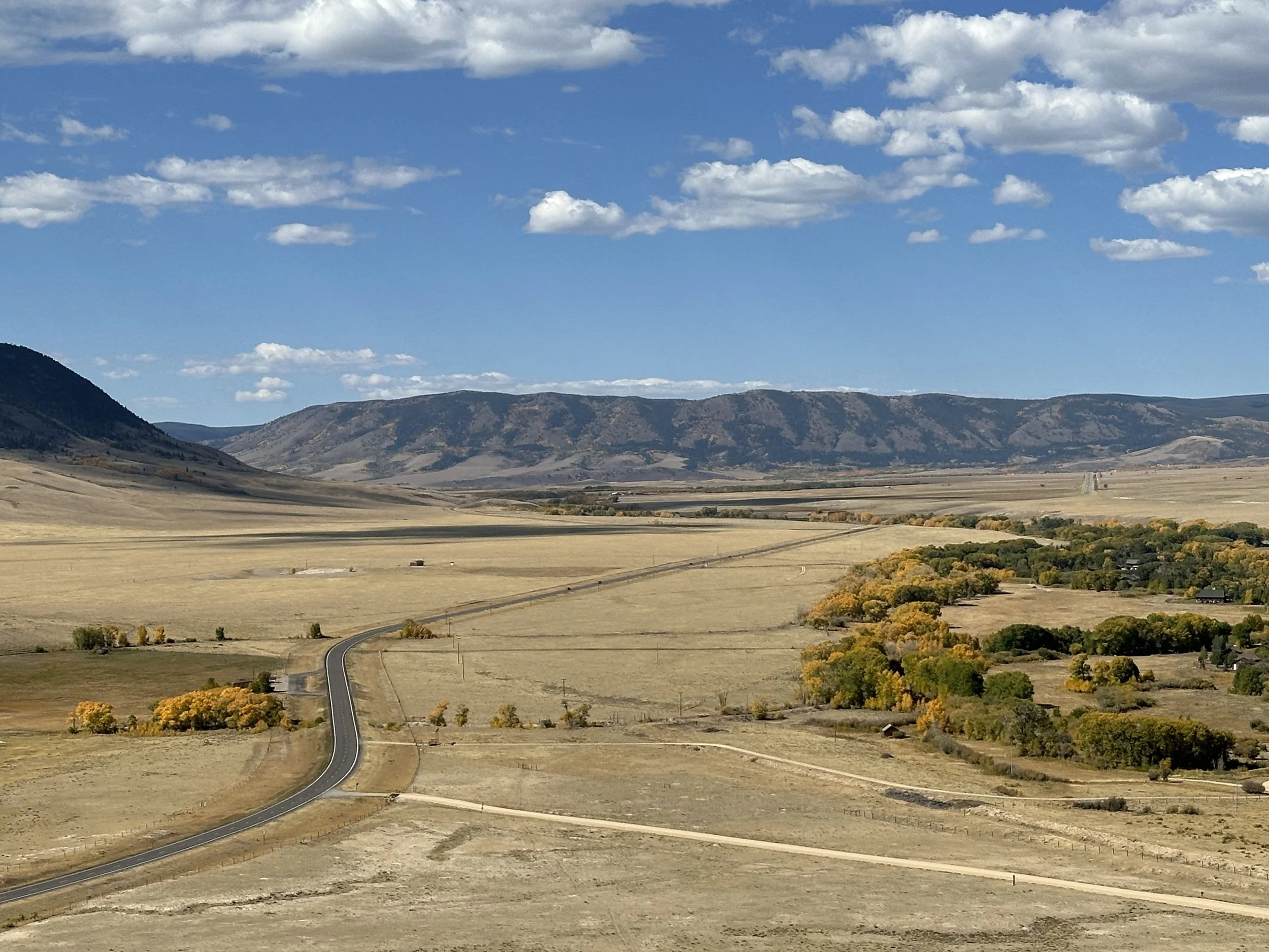 Open landscape with a winding road, yellow and green trees, fields, mountains in the background, and a partly cloudy sky, located near Cowgirl Publishing retreats location.