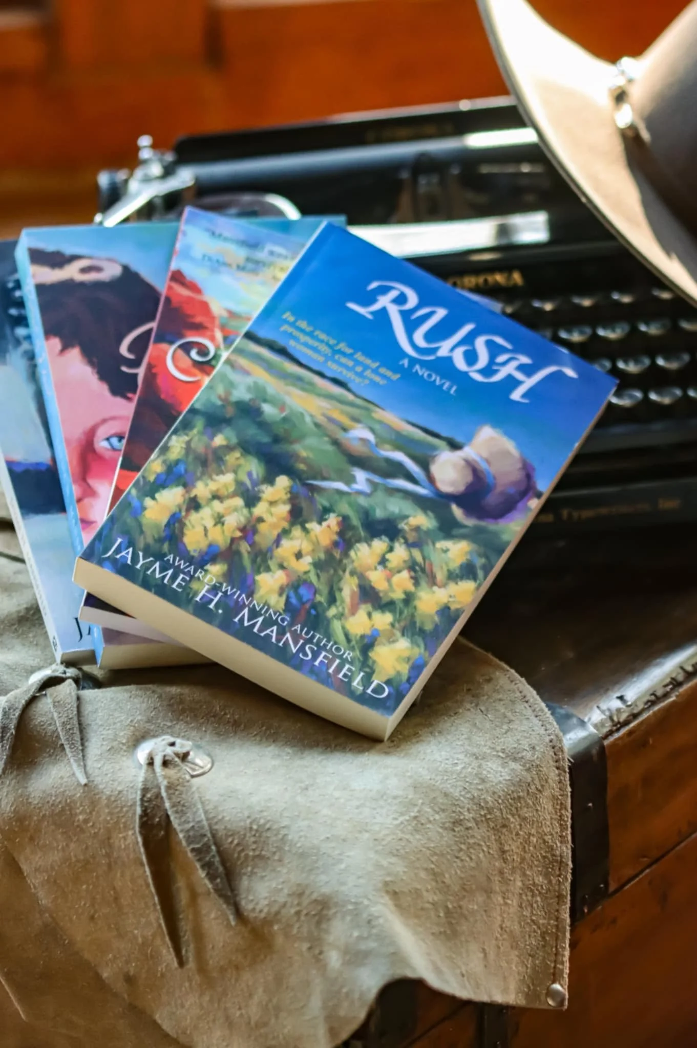 Three paperback books with colorful covers resting on a beige suede garment, along with a typewriter in the background. The book in the front is titled 'Rush' by Jayme H. Mansfield.
