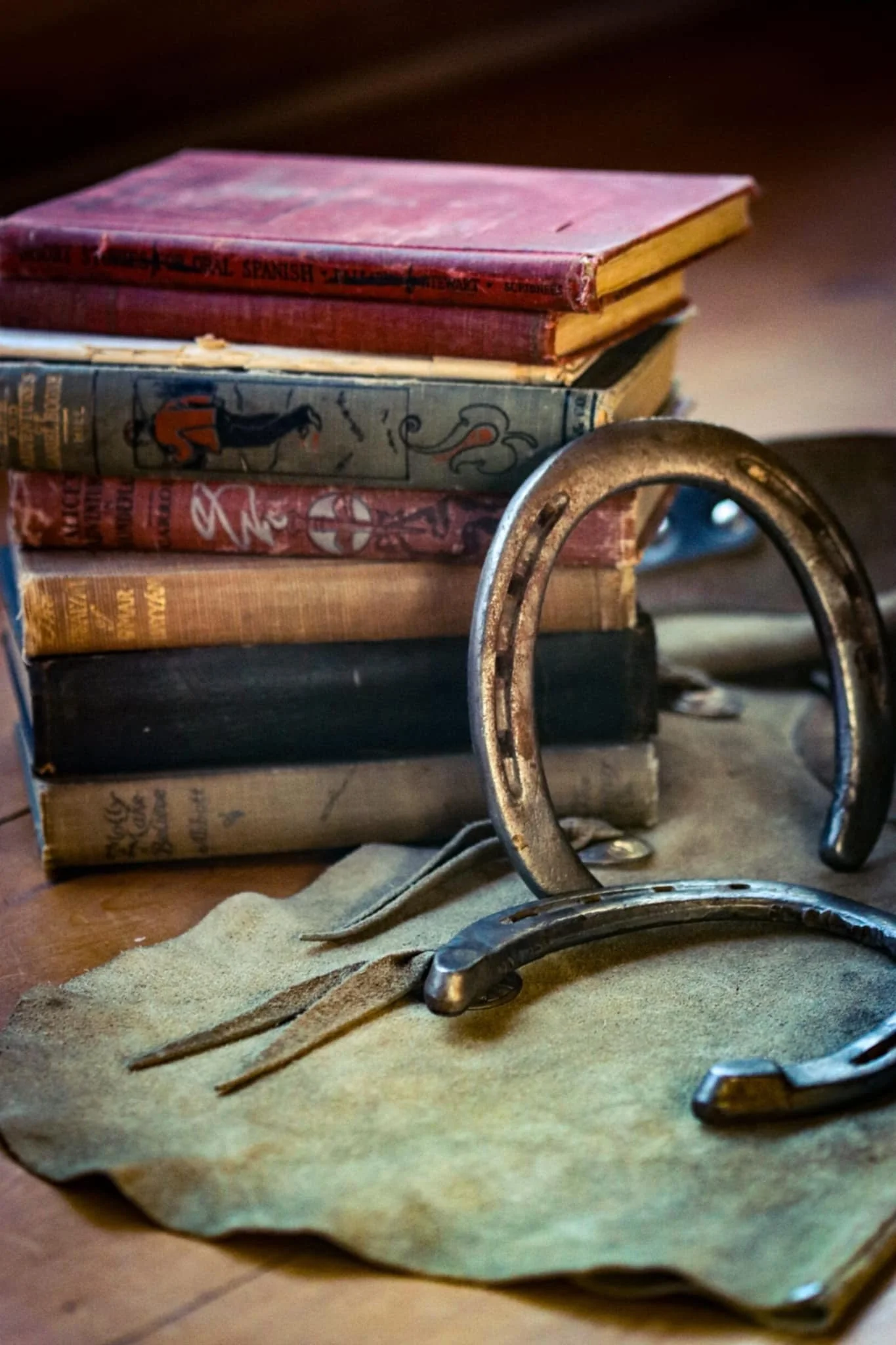 Stack of worn books, a horseshoe, and a leather strap on a piece of cloth on a wooden surface.
