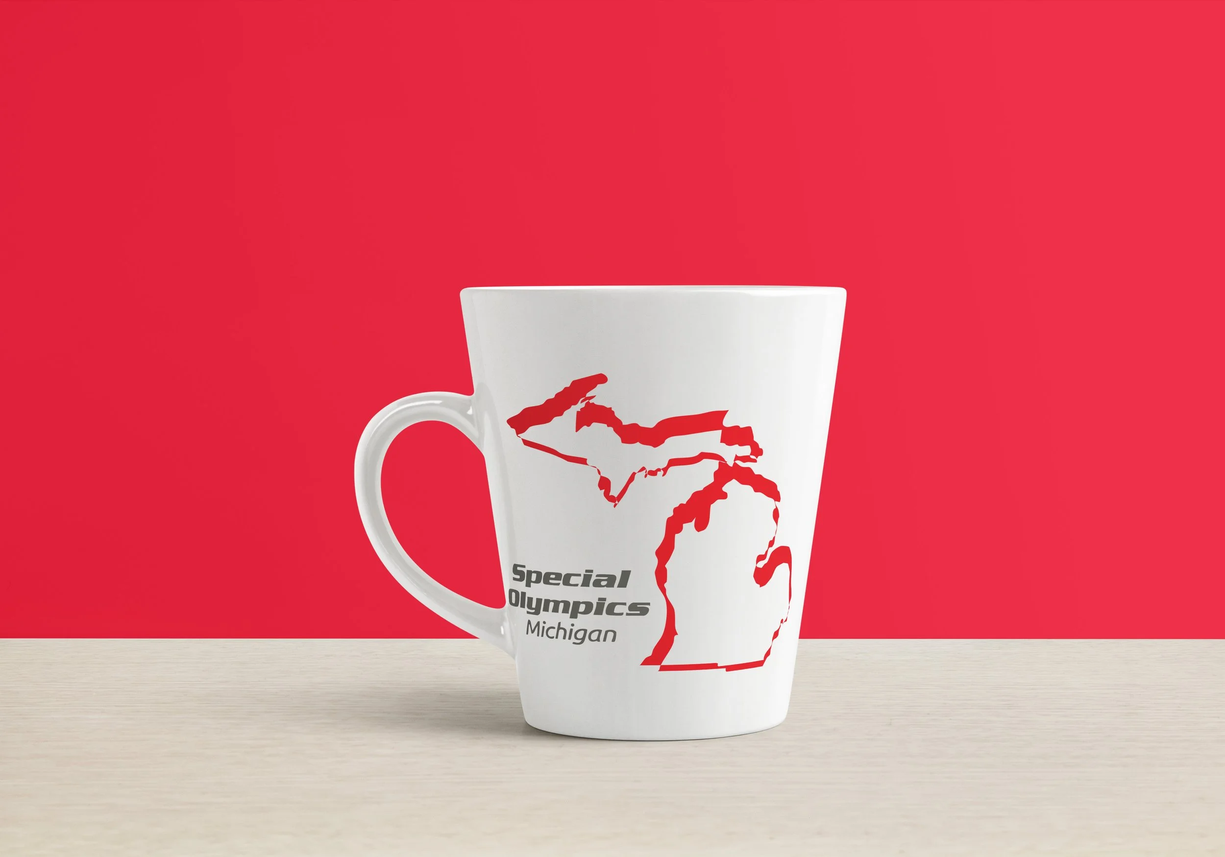 White mug with a red outline map of Michigan and the words "Special Olympics Michigan" on a beige surface against a red background.
