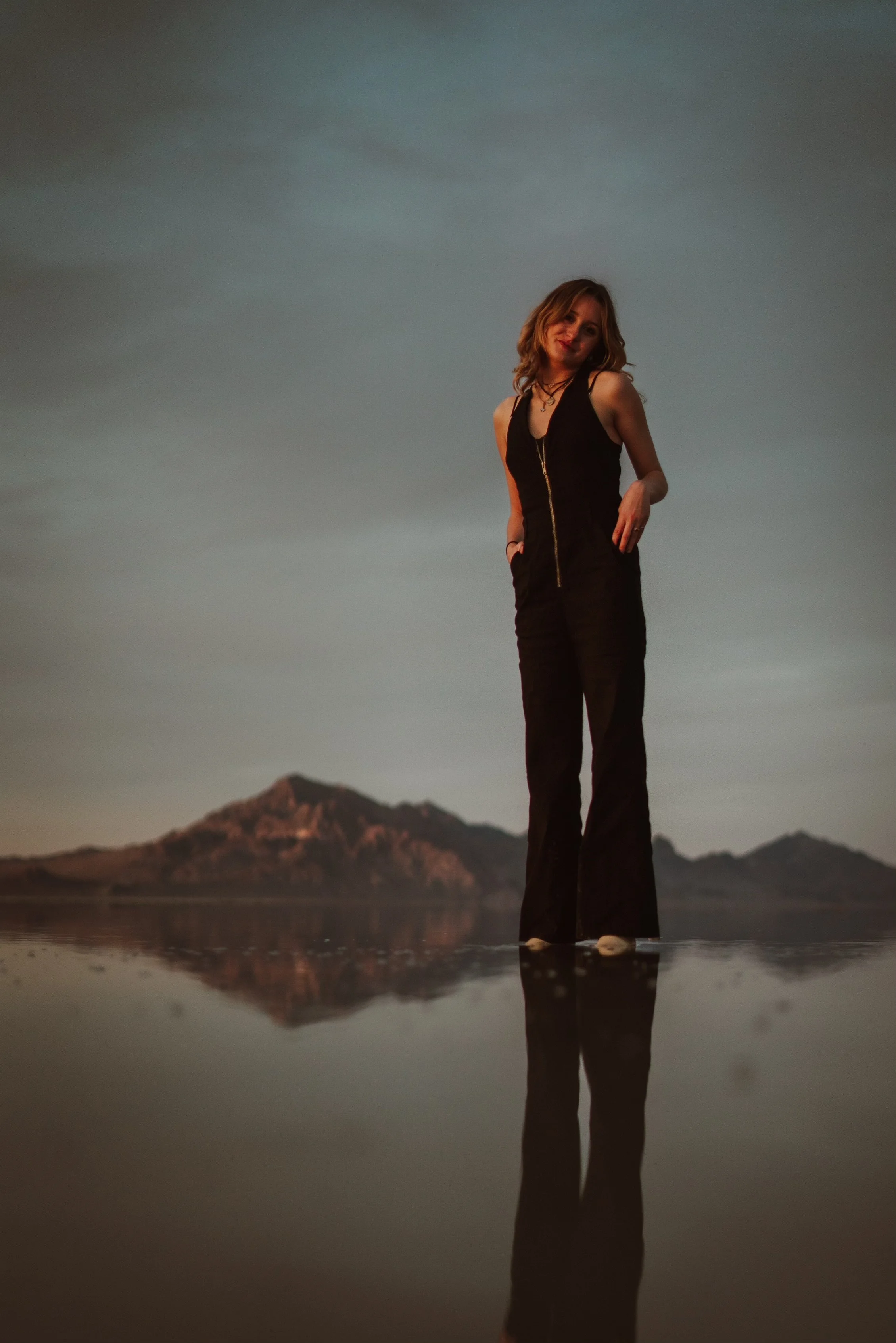 A woman standing on a reflective surface with mountains in the background during sunset or sunrise.