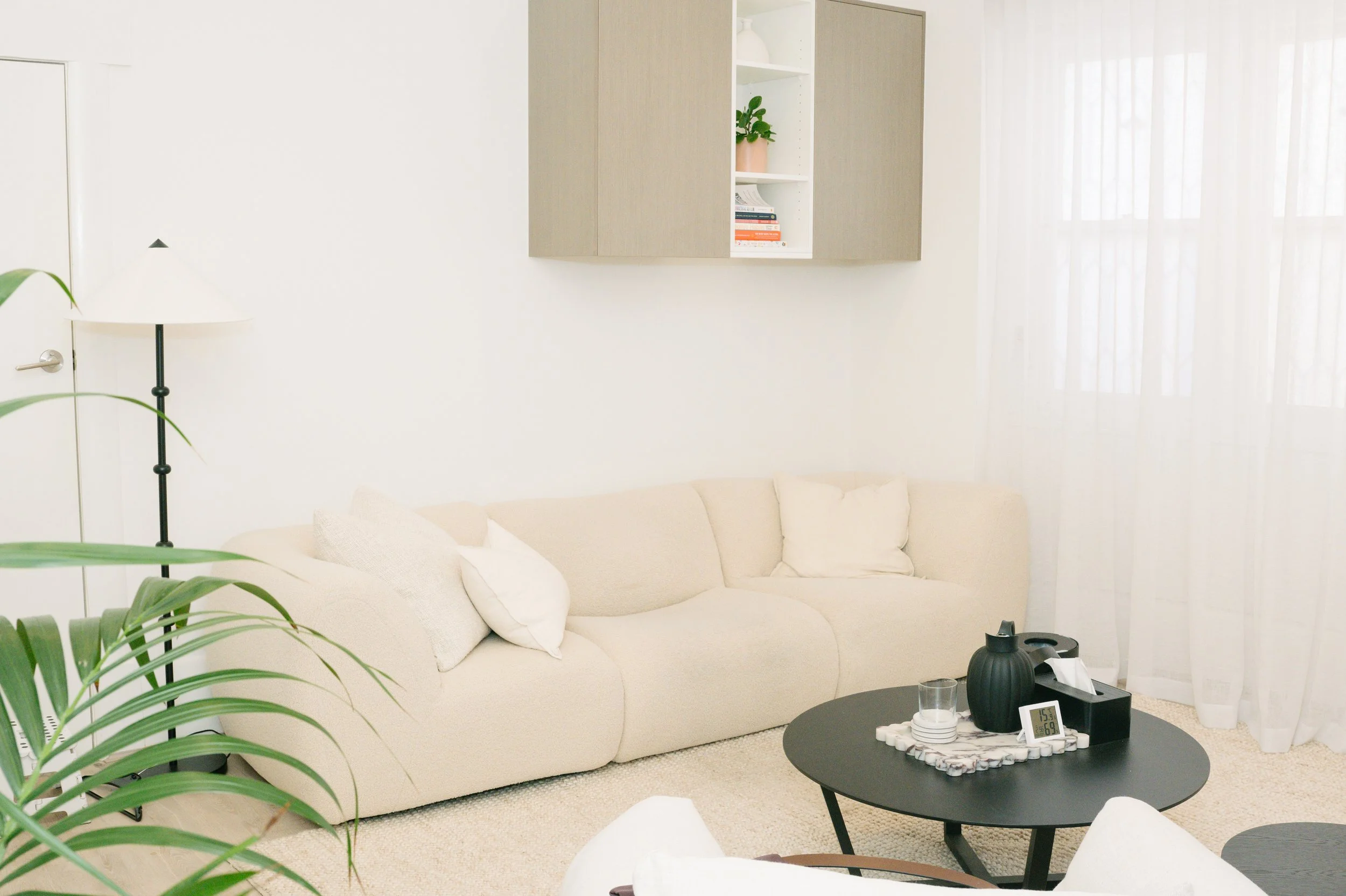 Consulting room with a cream sofa, black coffee table with a tray, plants, and a wall-mounted shelf with books and a small potted plant, sunlight coming through sheer curtains.