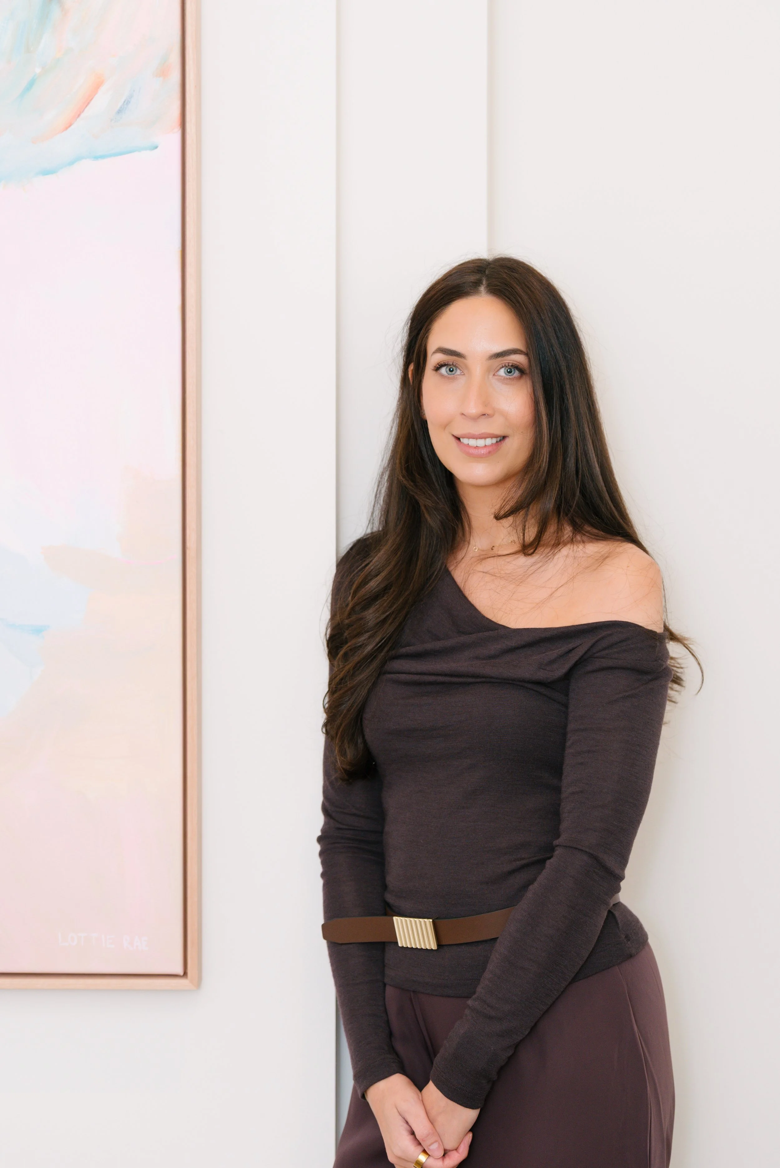 A young woman with long dark hair, blue eyes, and fair skin, smiling and standing against a white wall with a colorful abstract painting nearby.