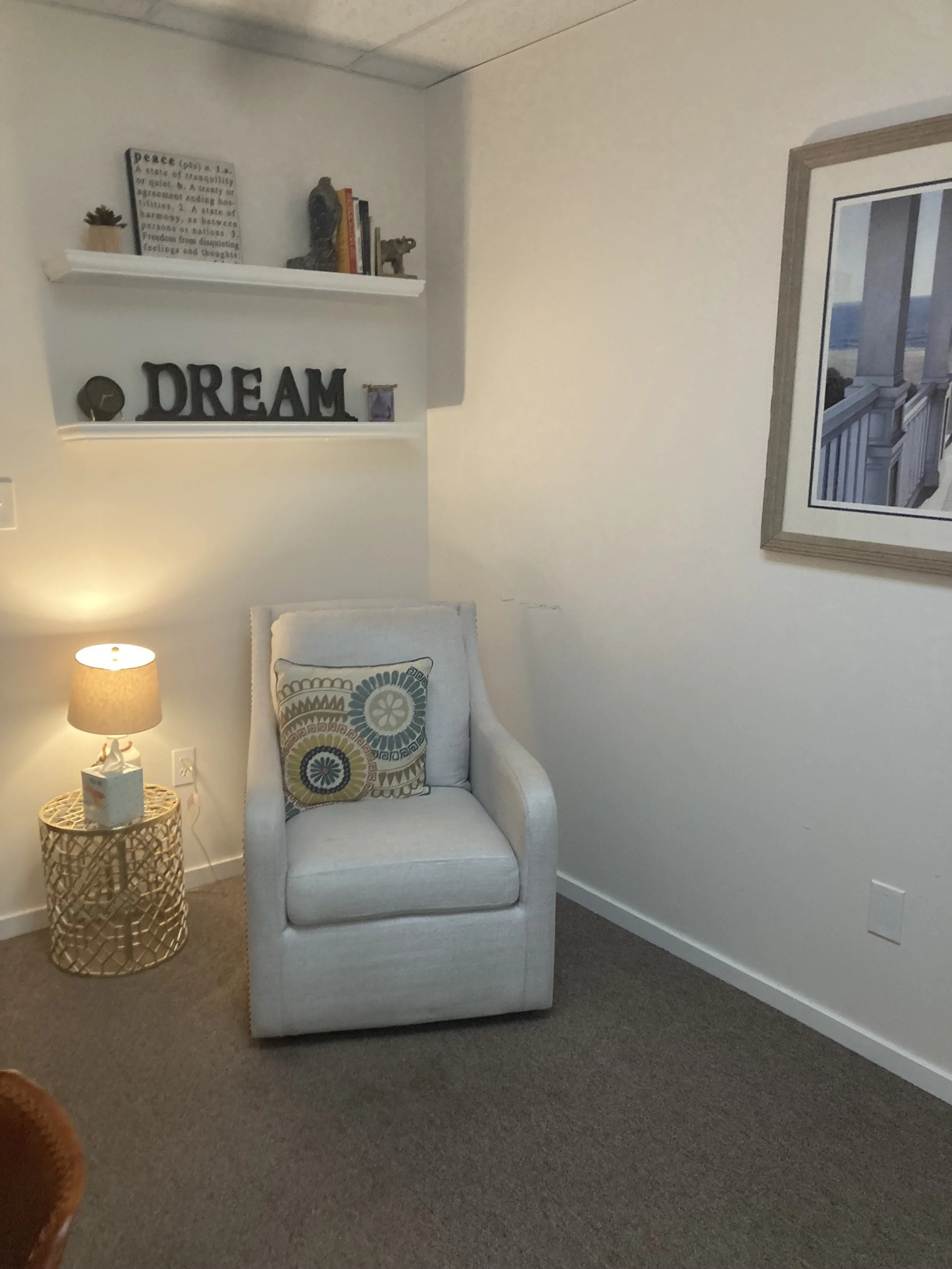 A cozy reading nook with a white armchair, a decorative pillow, a beige side table with a lattice design, a lit table lamp, floating shelves with books and decorative items, and a framed picture on the wall.