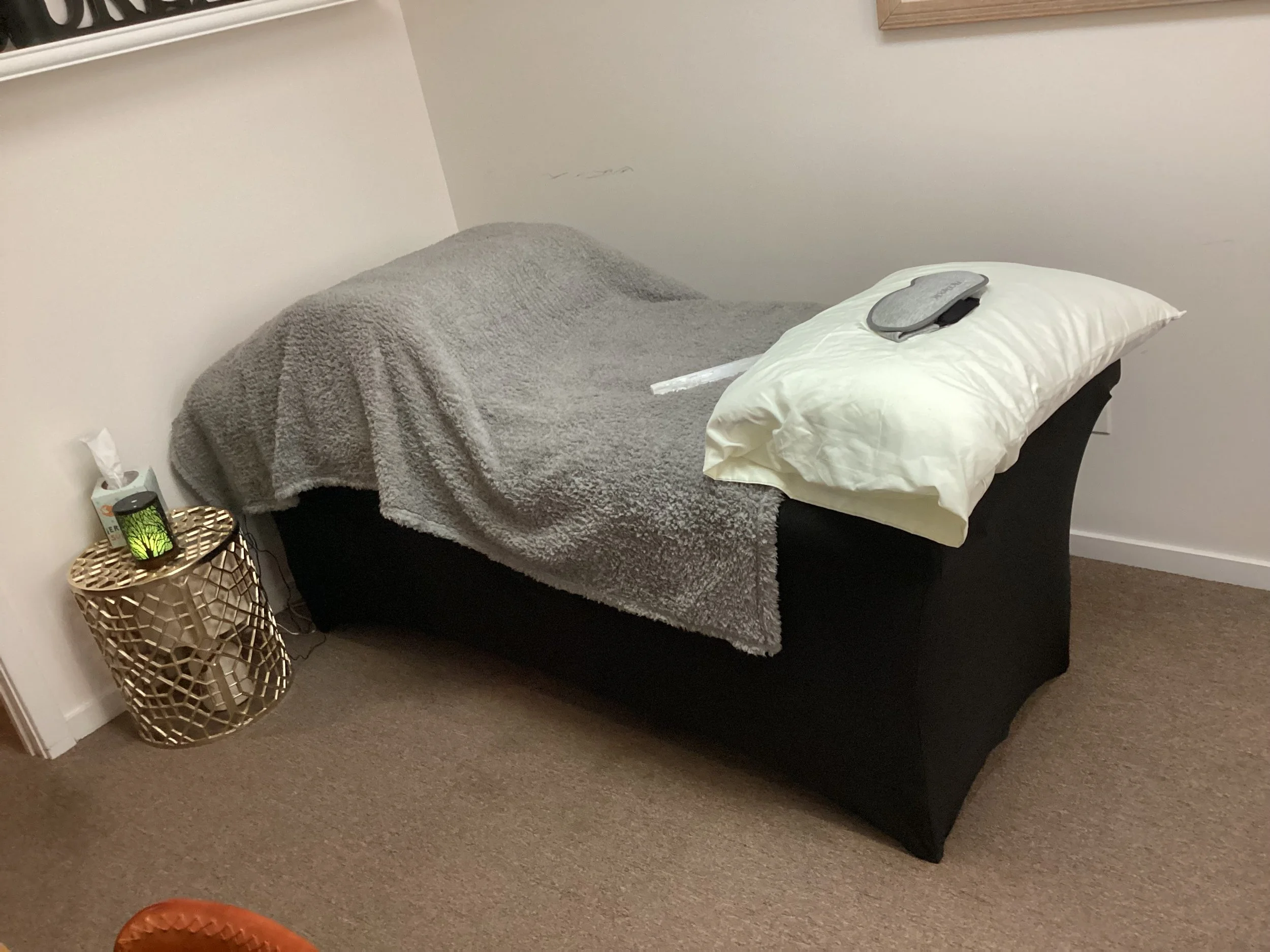 A massage or therapy bed covered with a gray blanket, with a white pillow and a gray eye mask on top, situated in a room with beige carpet and white walls.