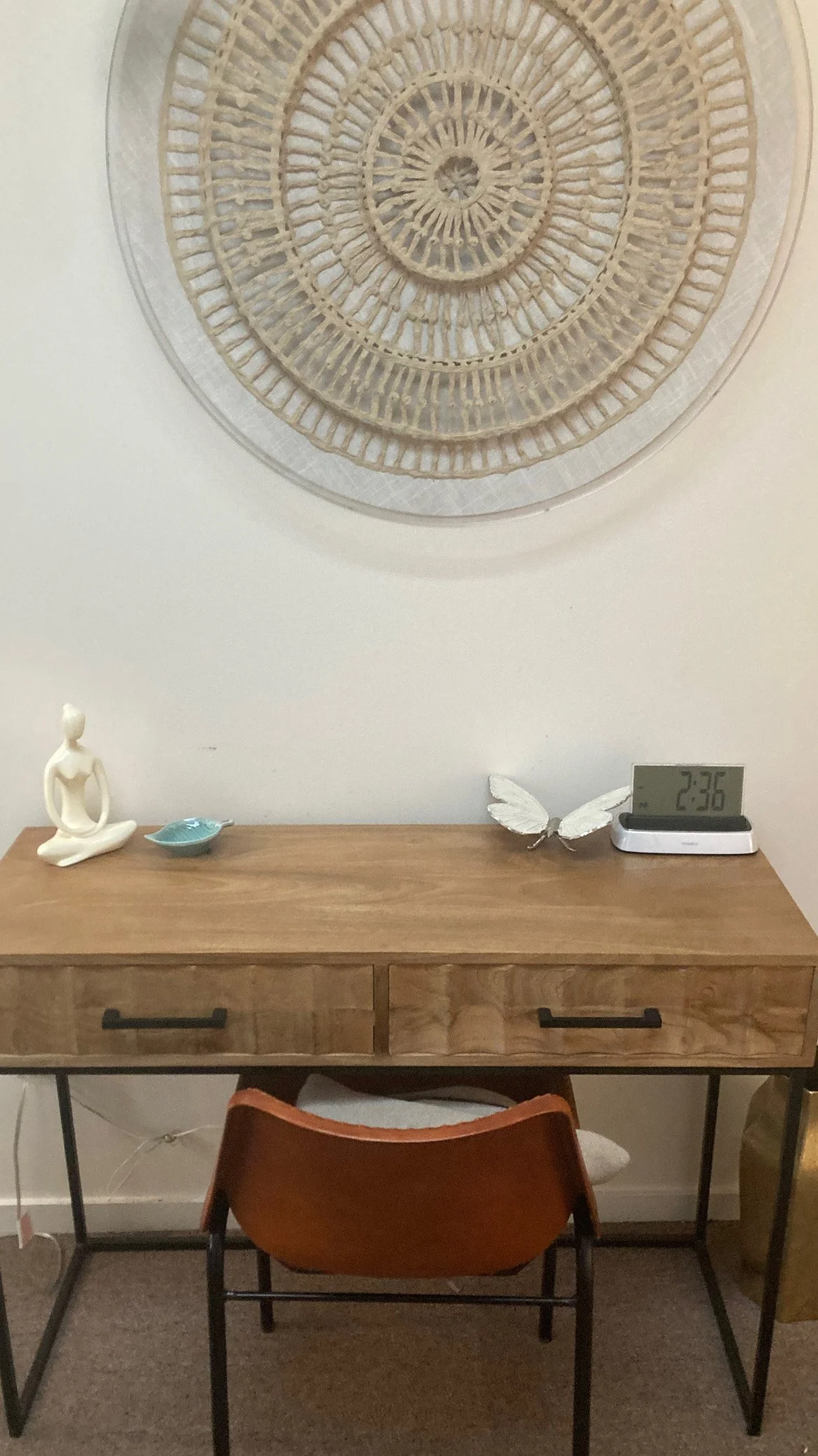A wooden desk with two drawers, a modern black-and-white digital clock showing 2:36, a ceramic sculpture of a seated woman meditating, a small blue dish, a white butterfly decorative piece, and a digital clock. The wall behind the desk has a large, c