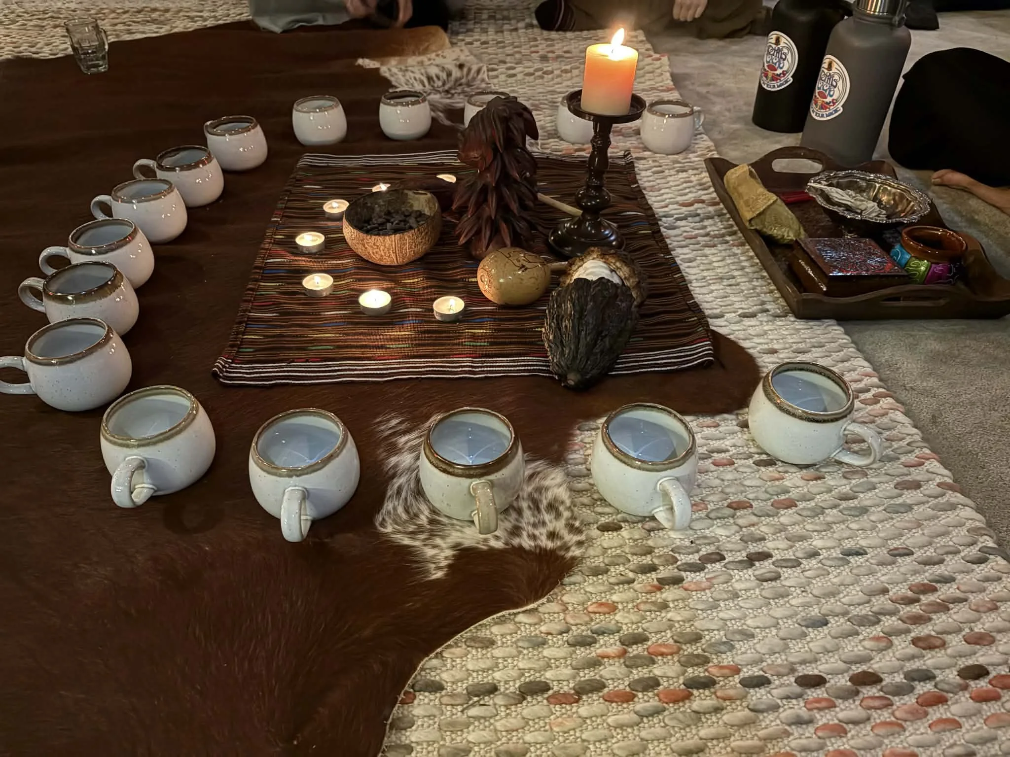 A table set with a central arrangement of candles, stones, and tribal objects, surrounded by white mugs with gold rims, and decorated with animal hides and woven fabric.