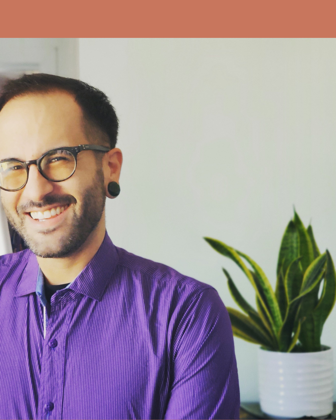 A man with dark hair, glasses, and a beard smiling at the camera, wearing a purple shirt, with a potted green plant in the background.