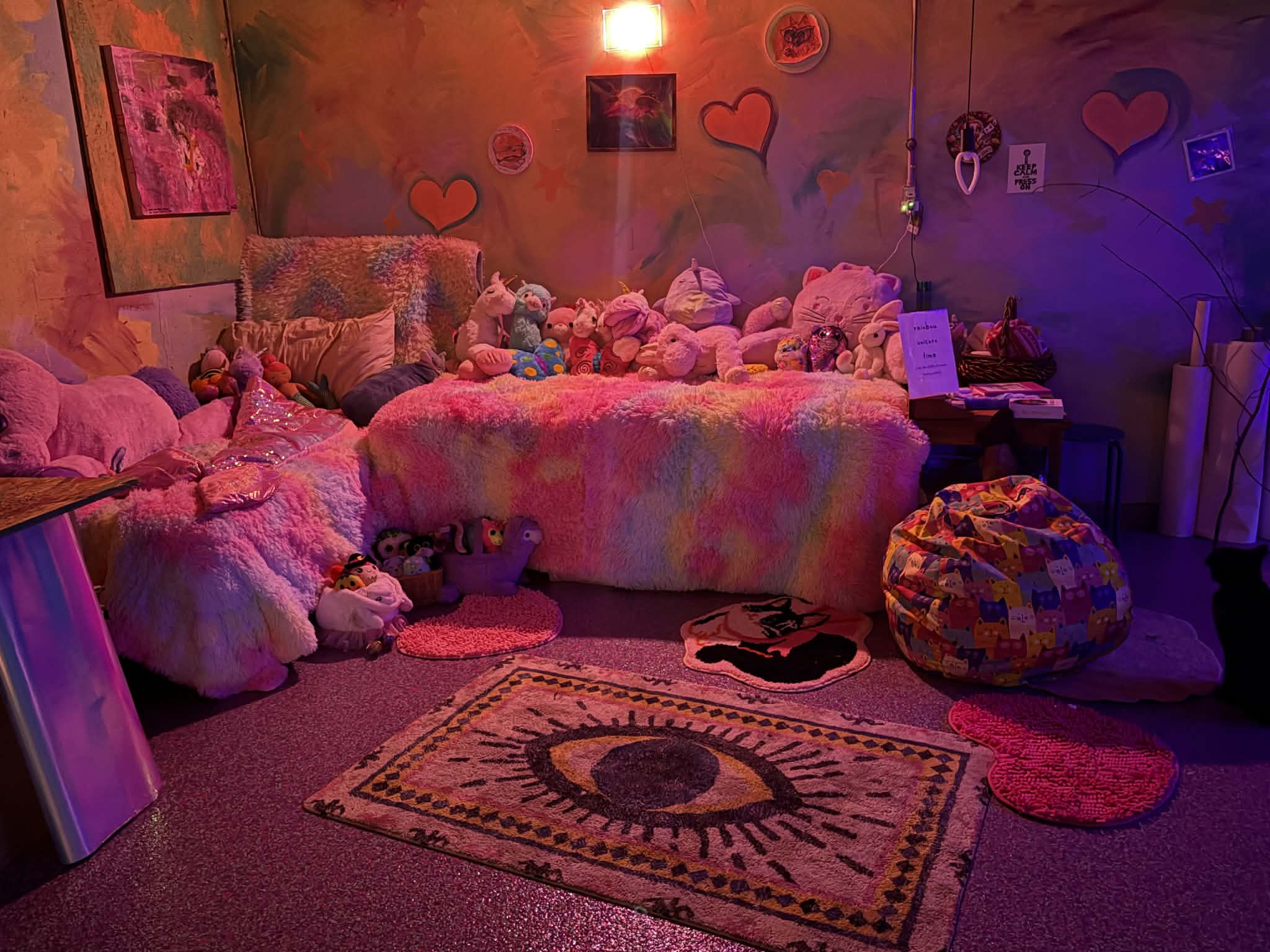 A cozy room decorated with plush toys and soft furnishings, including a bed or seating area covered with stuffed animals, pillows, and blankets. The walls are adorned with heart and star-shaped decorations, as well as framed pictures. The room is lit with pink and purple lighting, creating a warm and whimsical atmosphere.