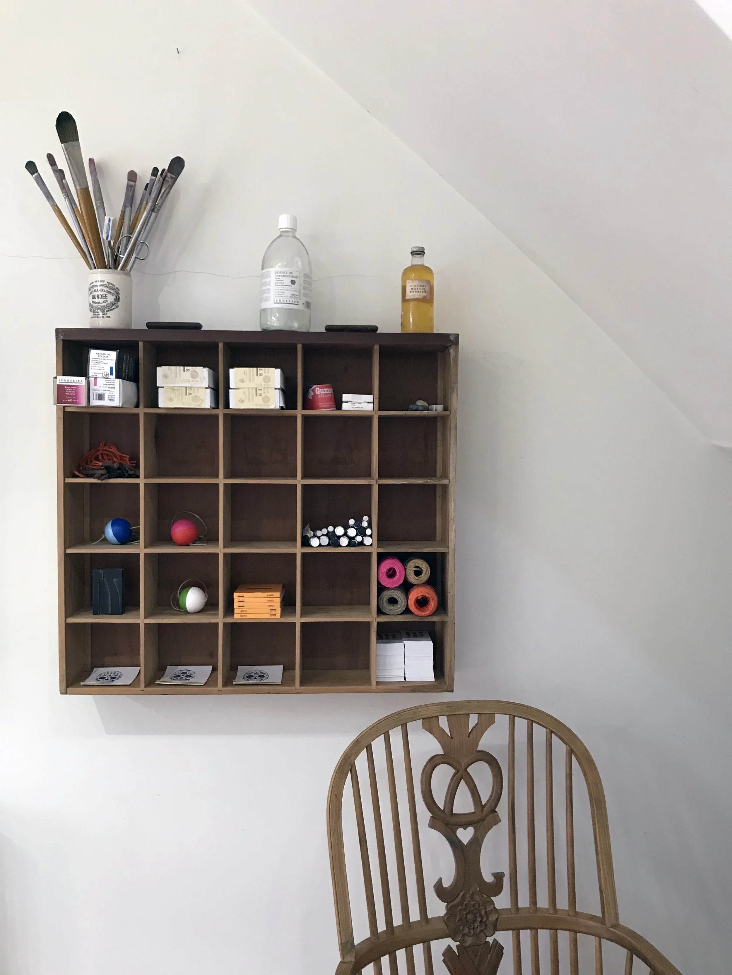 A wooden shelf mounted on a white wall holding bottles, boxes, and miscellaneous items, with a wooden chair partially visible in the foreground.