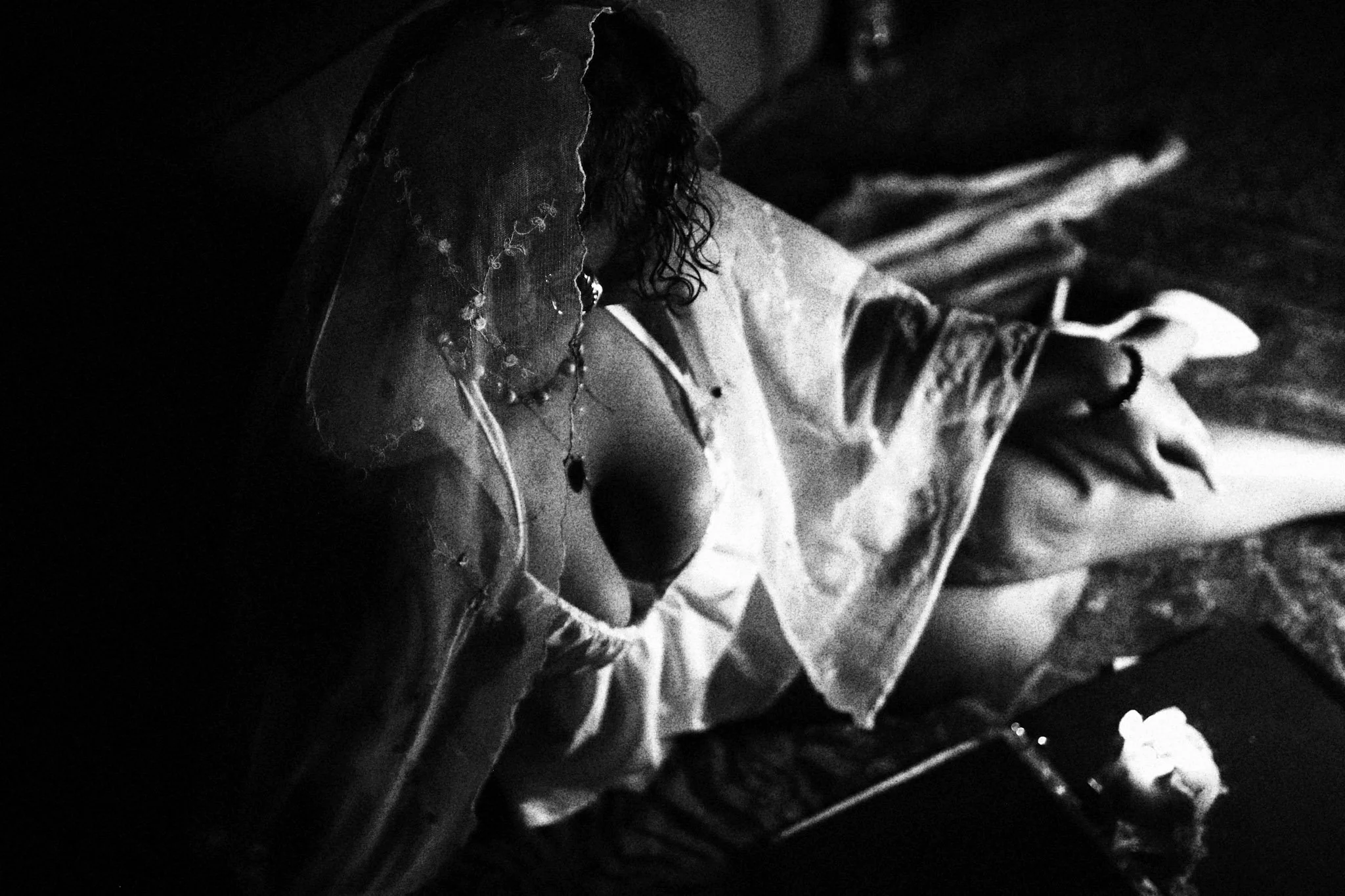 A woman with curly hair and layered necklaces is lying on her stomach on a bed, reading a book, with a phone and a lit candle nearby.