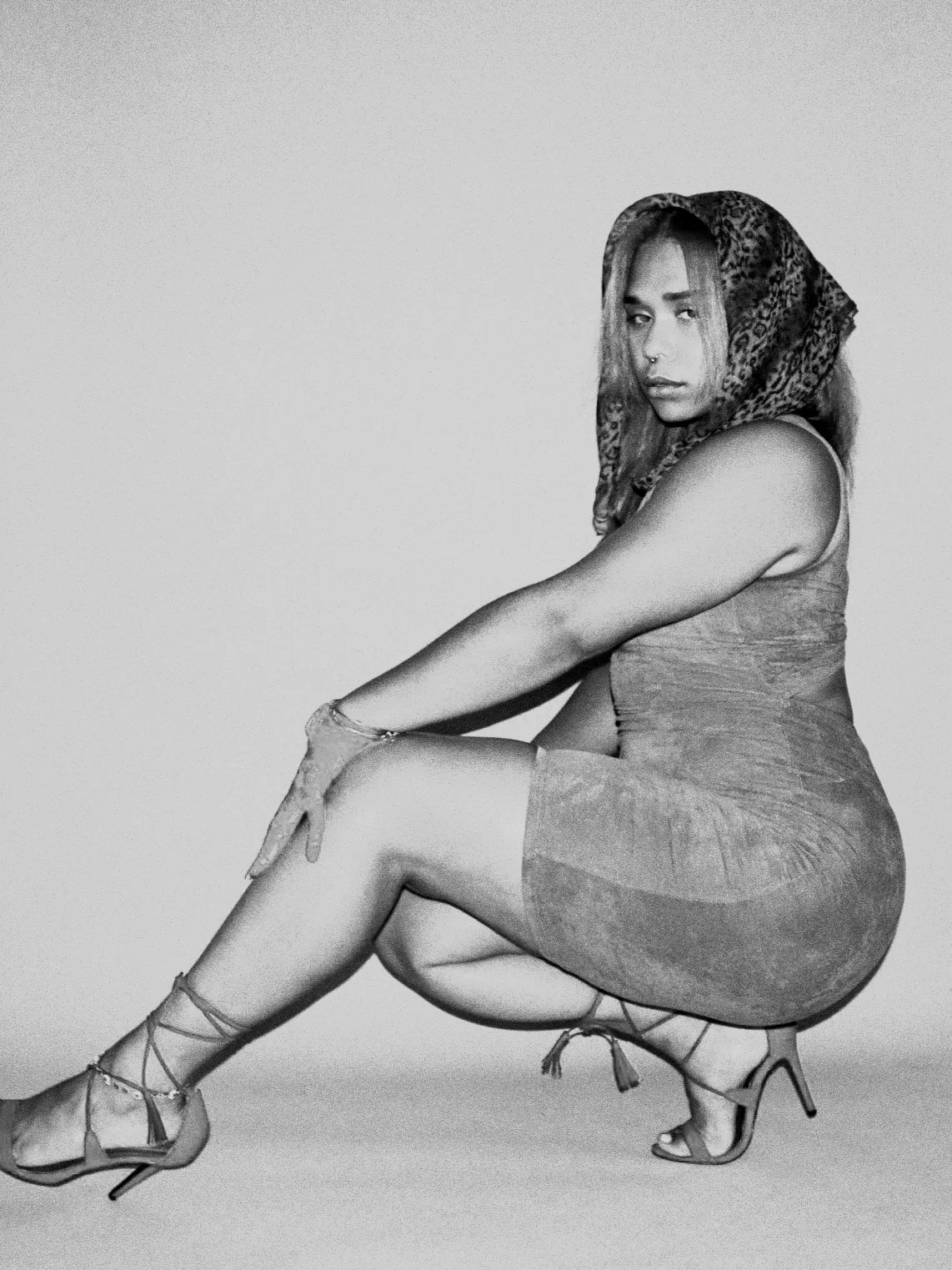 A woman crouching and posing for a photo, wearing a hooded headscarf, a sleeveless dress, and high heels.