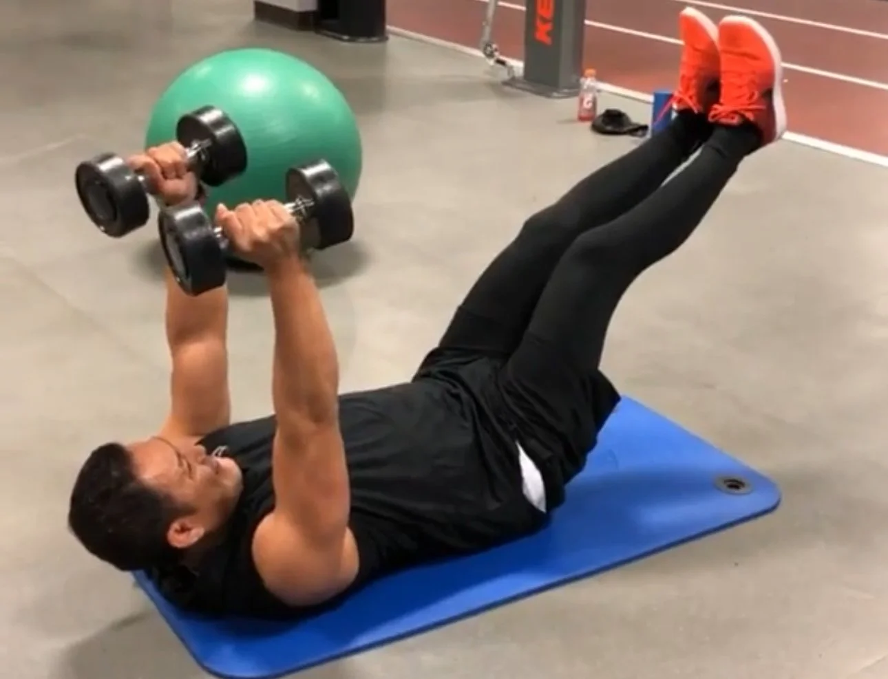 Man lying on blue exercise mat, doing a chest press with dumbbells in a gym, with a green exercise ball behind him and gym equipment in the background.