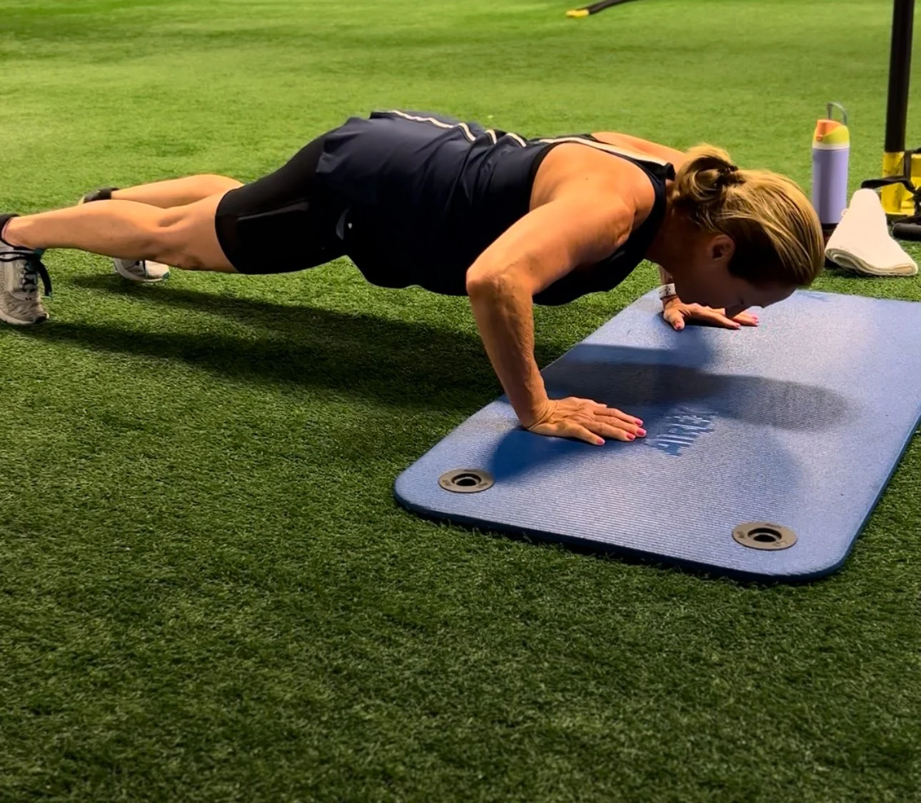 Woman doing a push-up on a blue exercise mat on artificial grass in a gym or fitness studio.