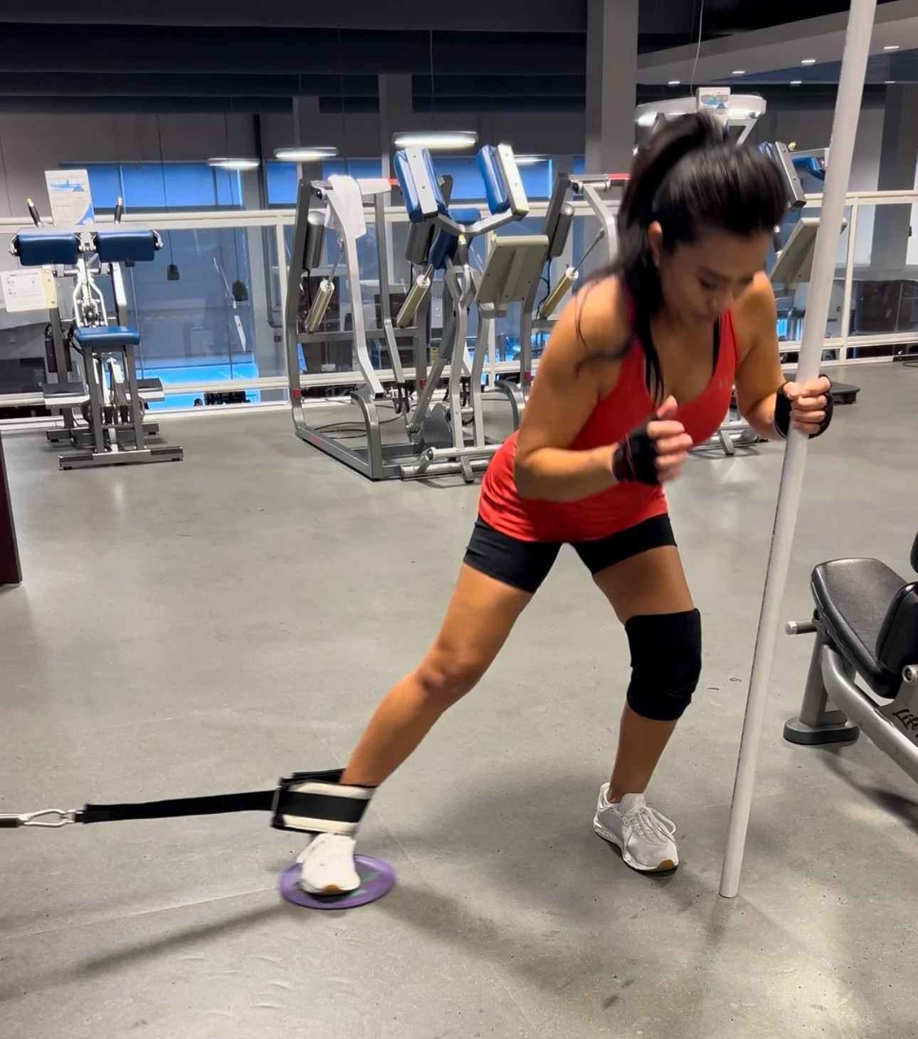 A woman wearing a red tank top, black shorts, and white sneakers training with a resistance band in a gym. She is holding a pole with one hand and leaning on a bench with the other.