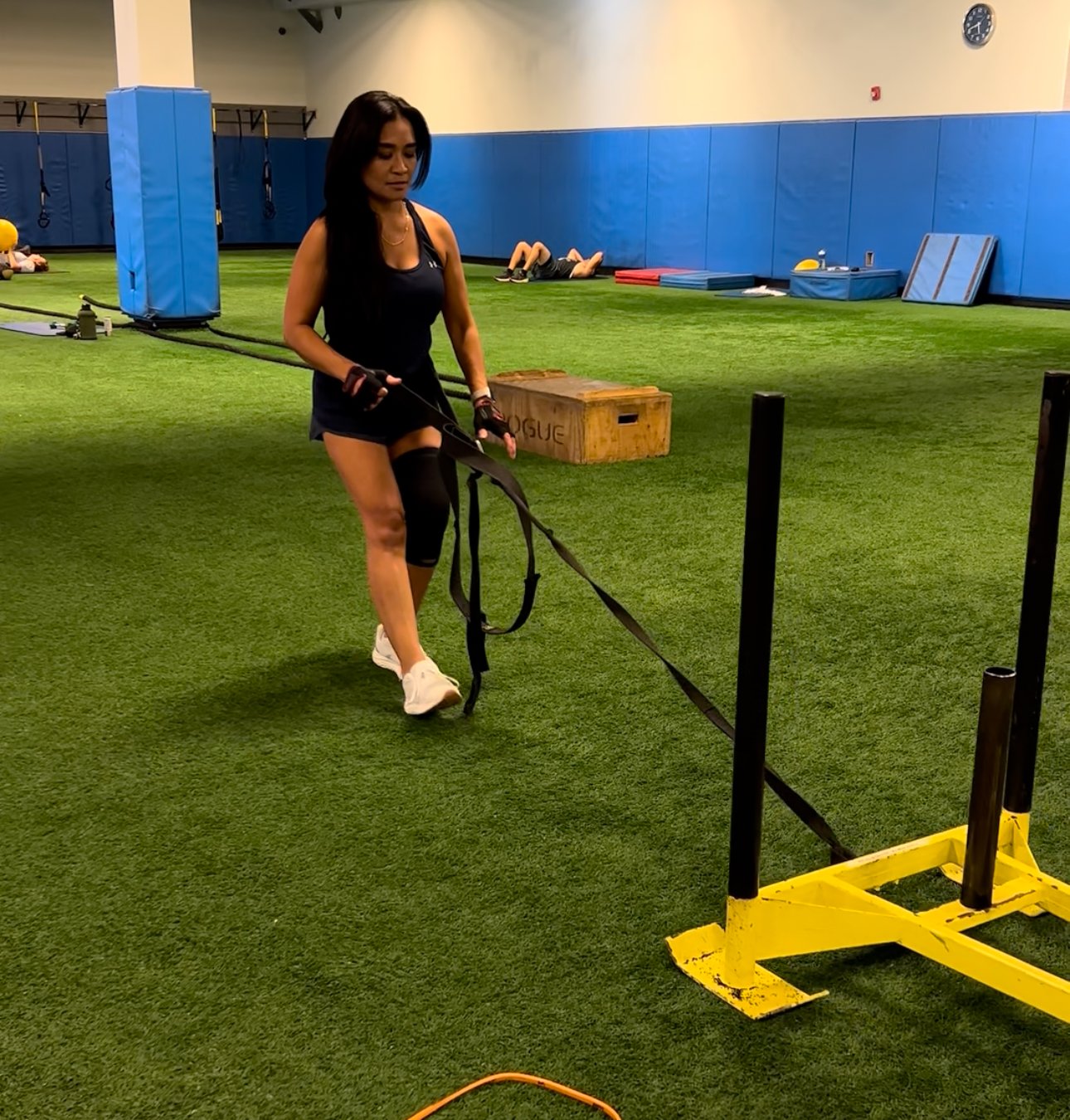 A woman in workout gear is walking indoors on green turf, pulling a resistance sled. She is wearing a black tank top, black shorts, white sneakers, and a knee brace. The gym has blue padded walls and various fitness equipment in the background.