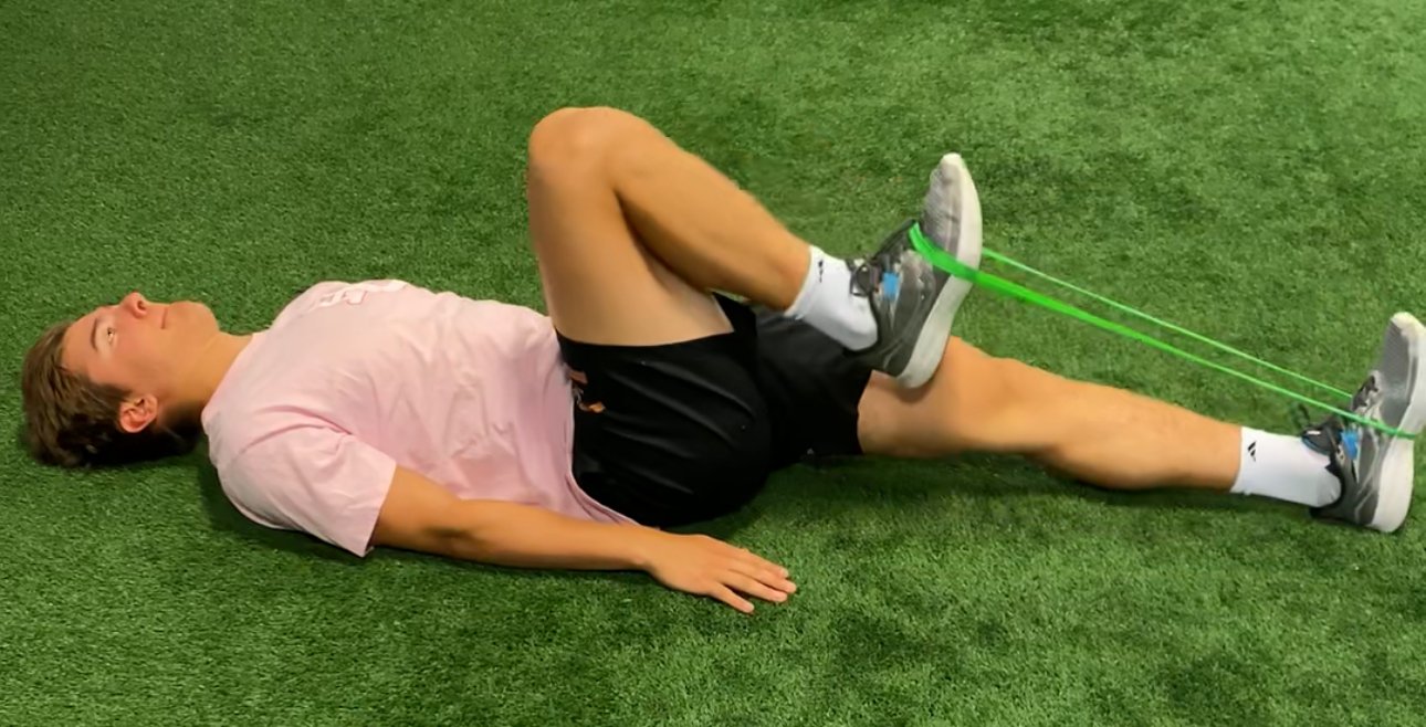 A man lying on his back on a green artificial turf, performing a leg stretch with resistance bands attached to his shoes.