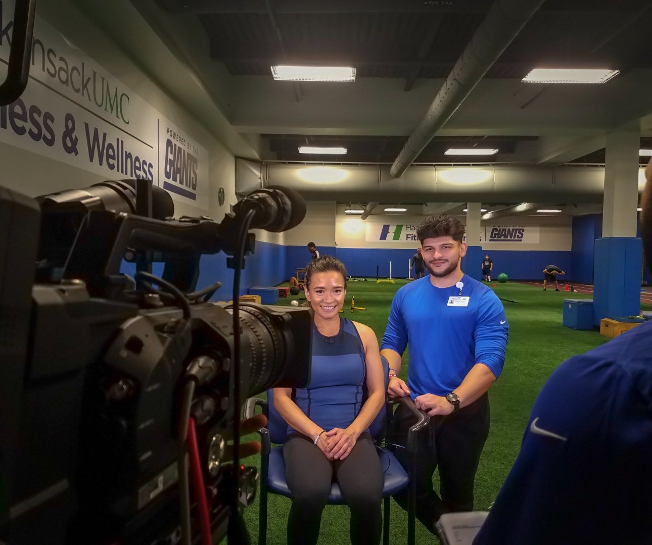 A woman sitting on a chair and a man standing beside her smiling, in an indoor gym or training facility with green turf and exercise equipment in the background, while a camera captures the scene.