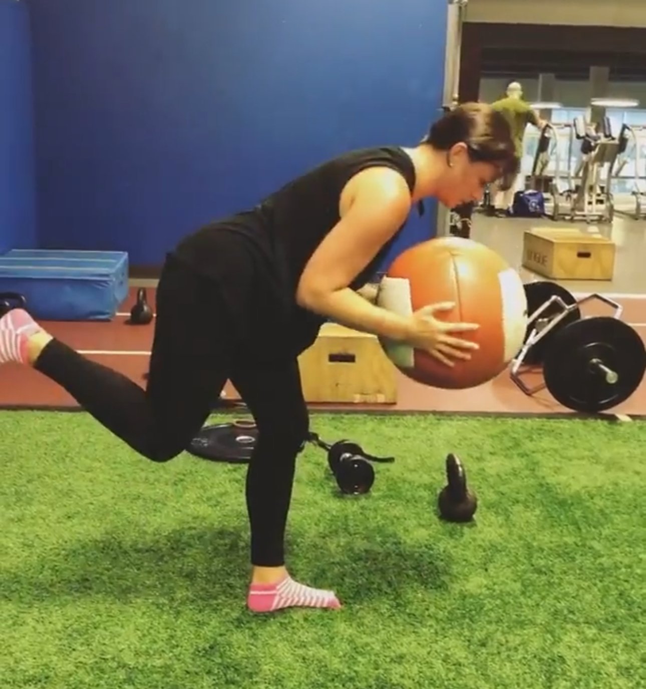 A woman in black workout clothes, wearing pink and white striped socks, is performing a fitness exercise on artificial grass, holding a large orange and white medicine ball. She is on one knee with her left leg extended behind her, in a gym setting.
