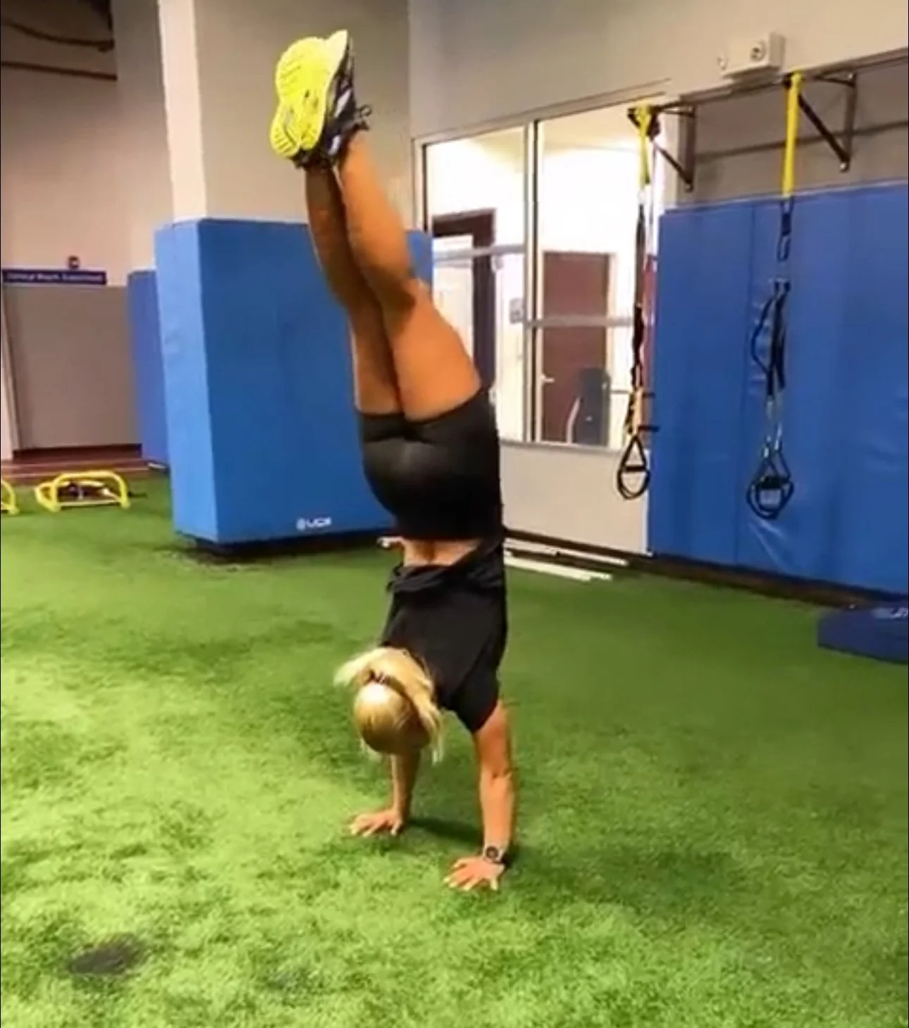 A woman performing a handstand in a gym, with her legs extended upward and wearing yellow sneakers.