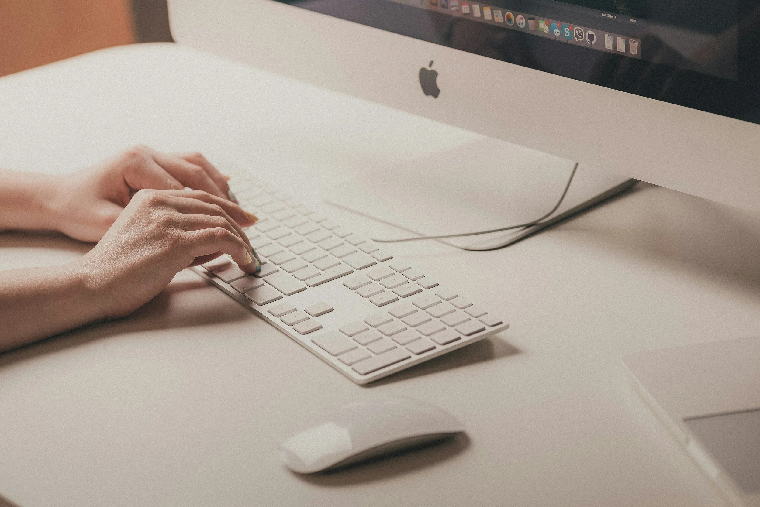 Hands typing on an Apple keyboard next to an iMac and a white mouse.