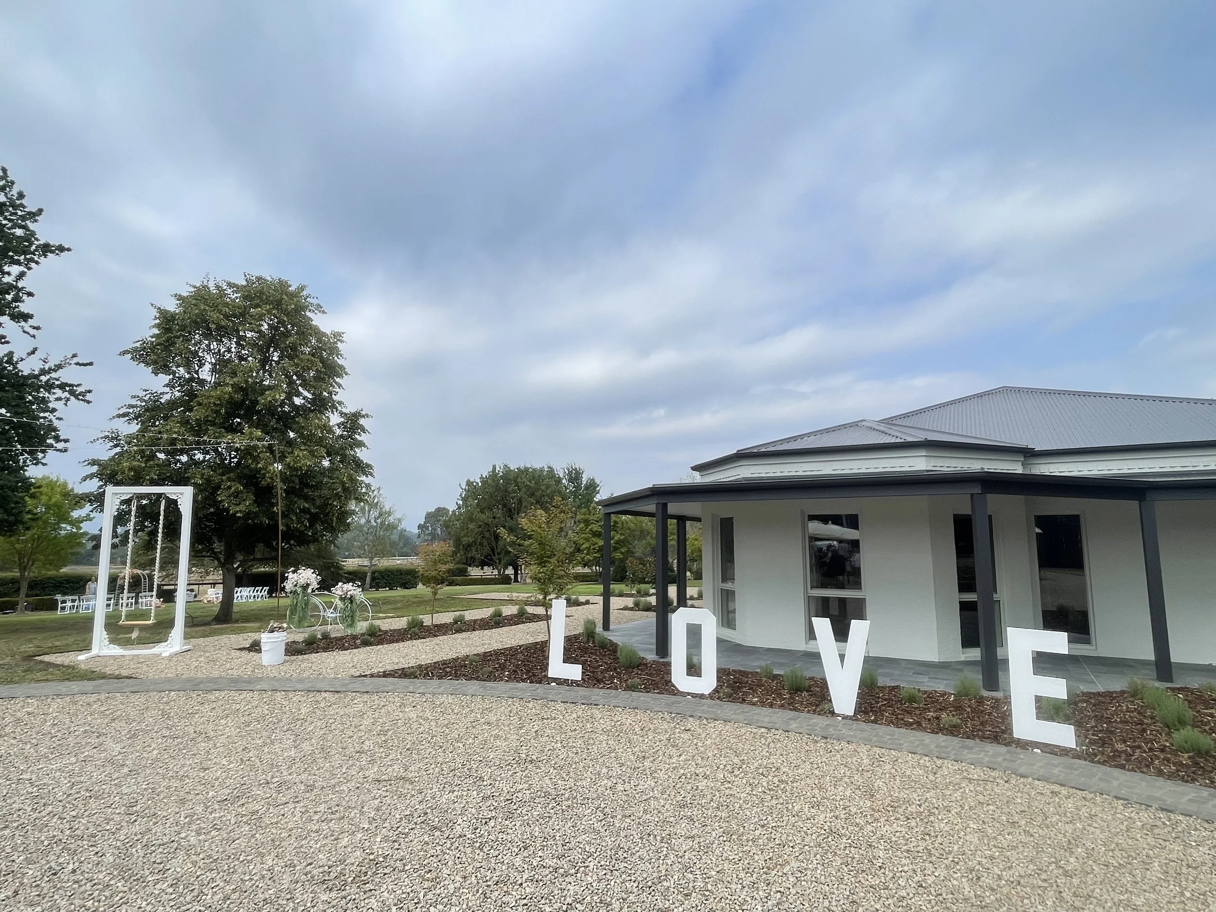 Outdoor wedding setup with large white decorative letters spelling "LOVE" in front of a white building with a gray roof and black trim. There is a white swing decorated with white flowers and green foliage on the left side, along with trees and a gra