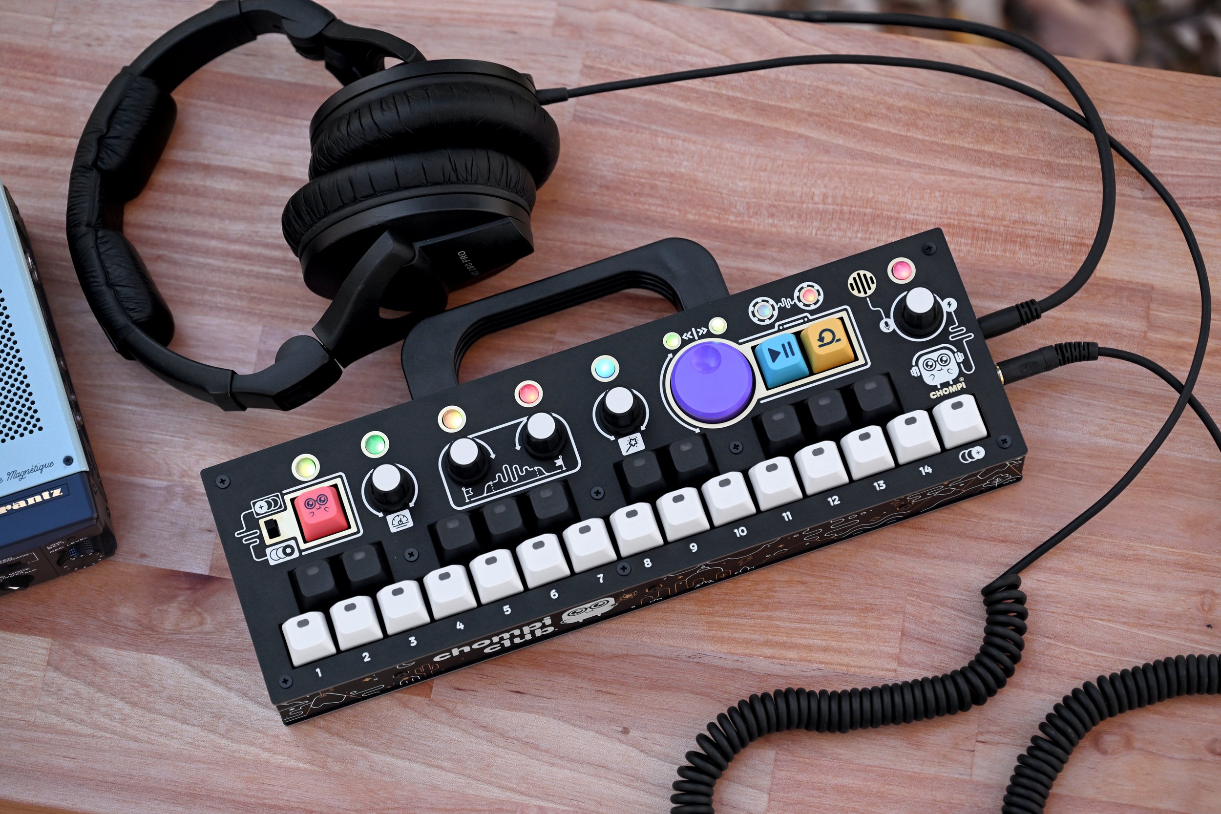 A compact electronic music production device with colorful knobs, buttons, and a small keyboard, placed on a wooden surface with headphones nearby.
