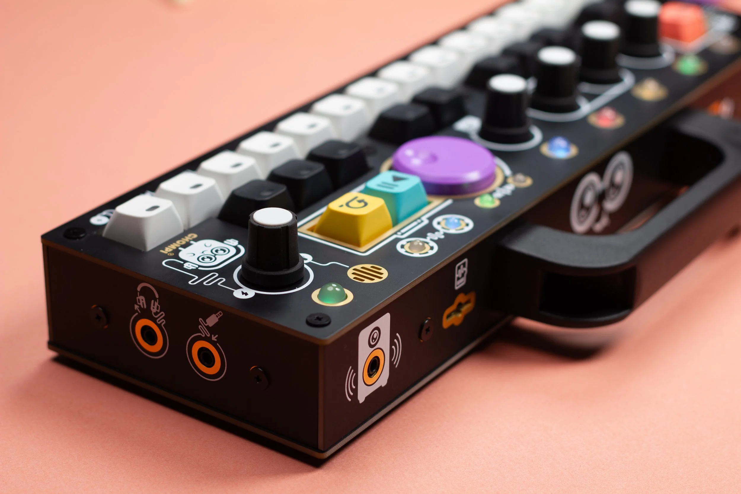 A back angle view of the Classic Black CHOMPI Sampler, a compact electronic music sampler with colorful buttons, knobs, and icons on a pink background. Showing the Master Line Output, Micro SD Card slot, CHOMPI handle, and surface controls.