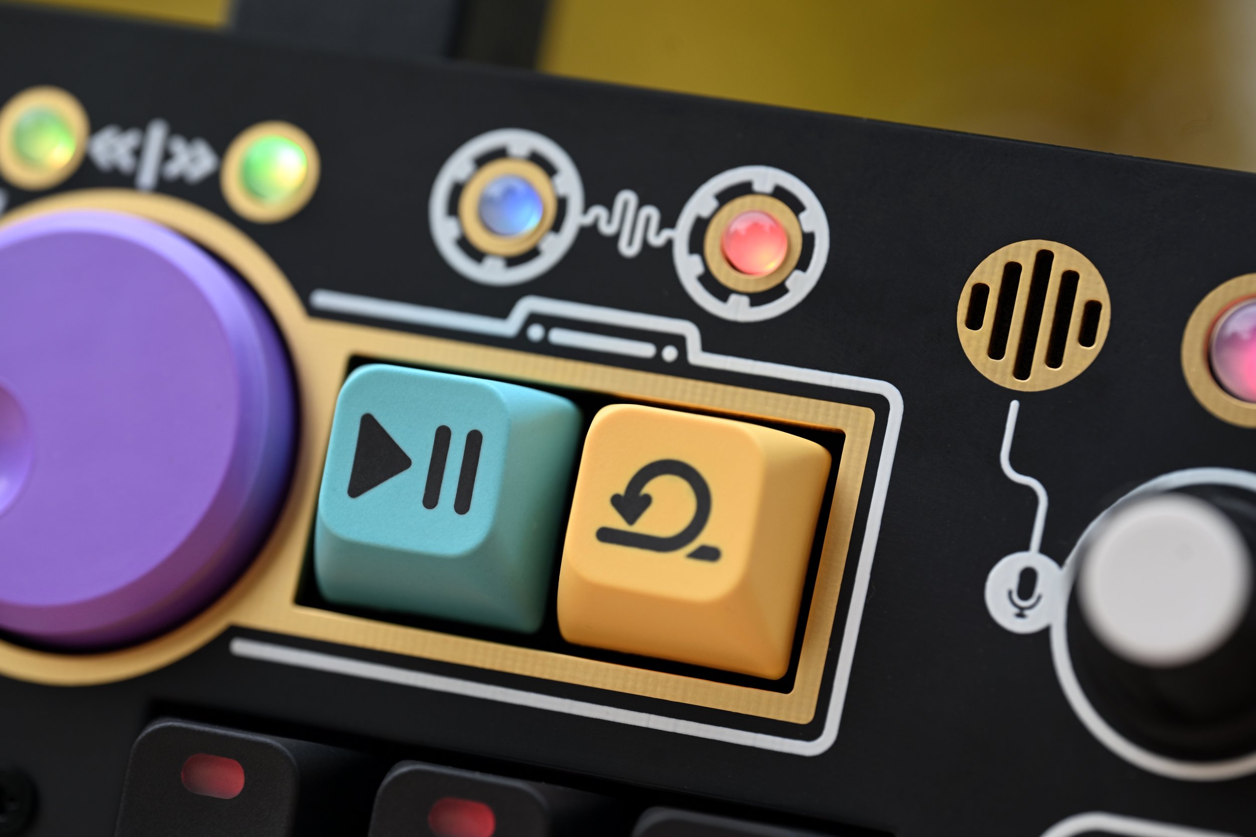 Close-up photo of a colorful electronic device featuring two buttons with play/pause and rewind icons, surrounded by various illuminated indicator lights and design elements.