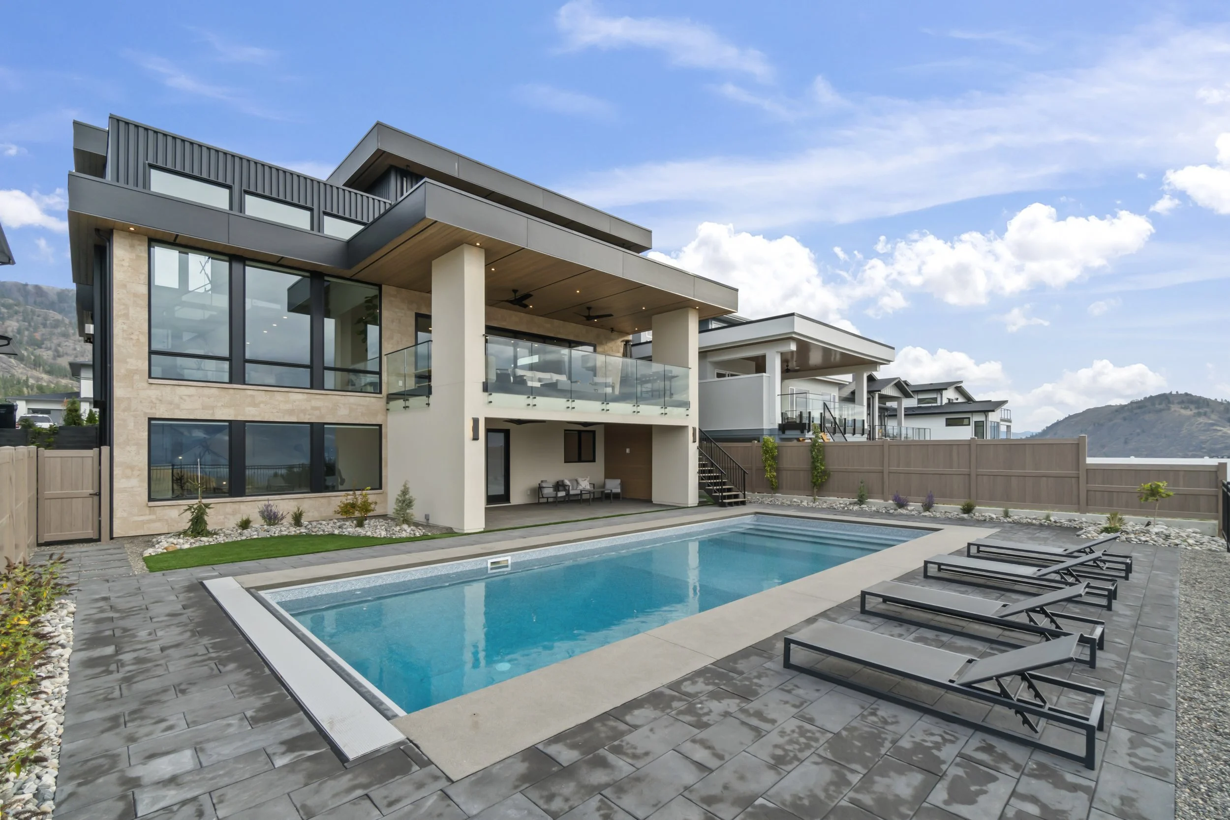 Modern house with large glass windows and an outdoor swimming pool, surrounded by lounge chairs and a wooden fence, with a mountainous landscape and blue sky in the background.