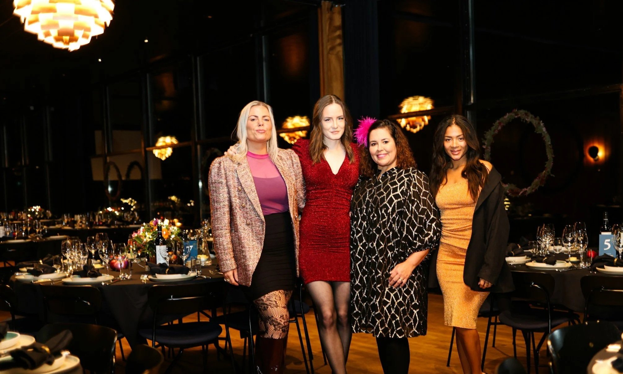 Four women standing together at a banquet table in a dimly lit, elegant event venue with round tables, wine glasses, and floral centerpieces