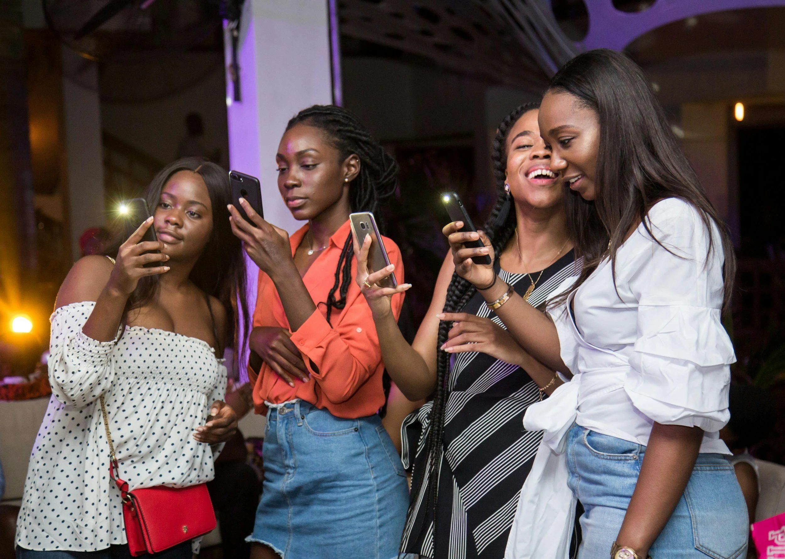 Group of five women at an indoor event, looking at their smartphones, with one woman smiling and hugging another.