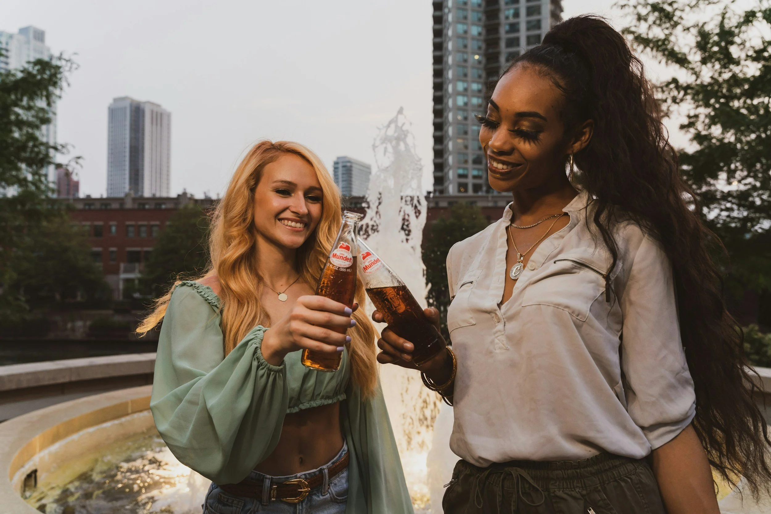 Two women smiling and toasting with bottled drinks in front of a fountain with city buildings in the background.
