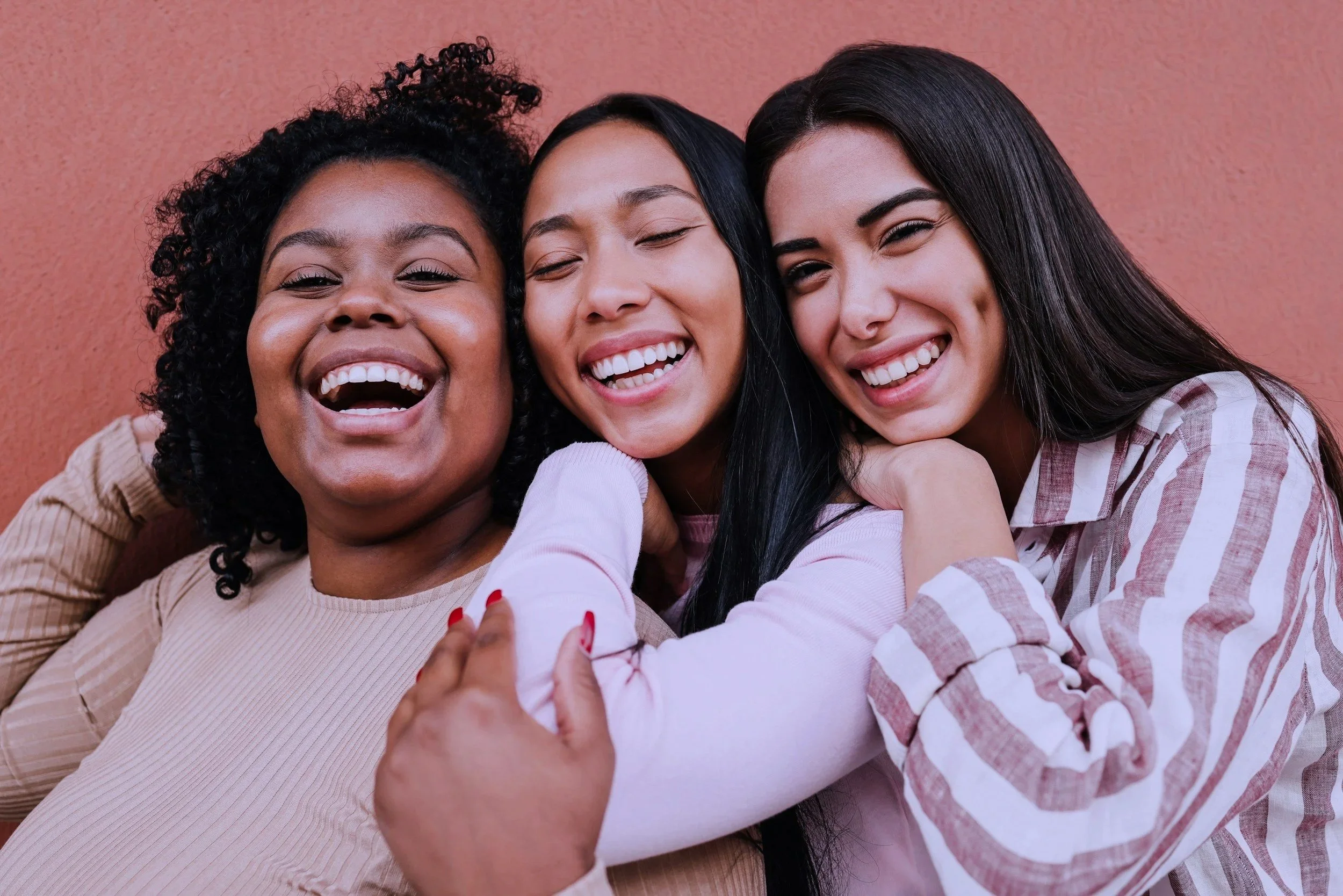 Three women smiling and hugging close together against a pink background.