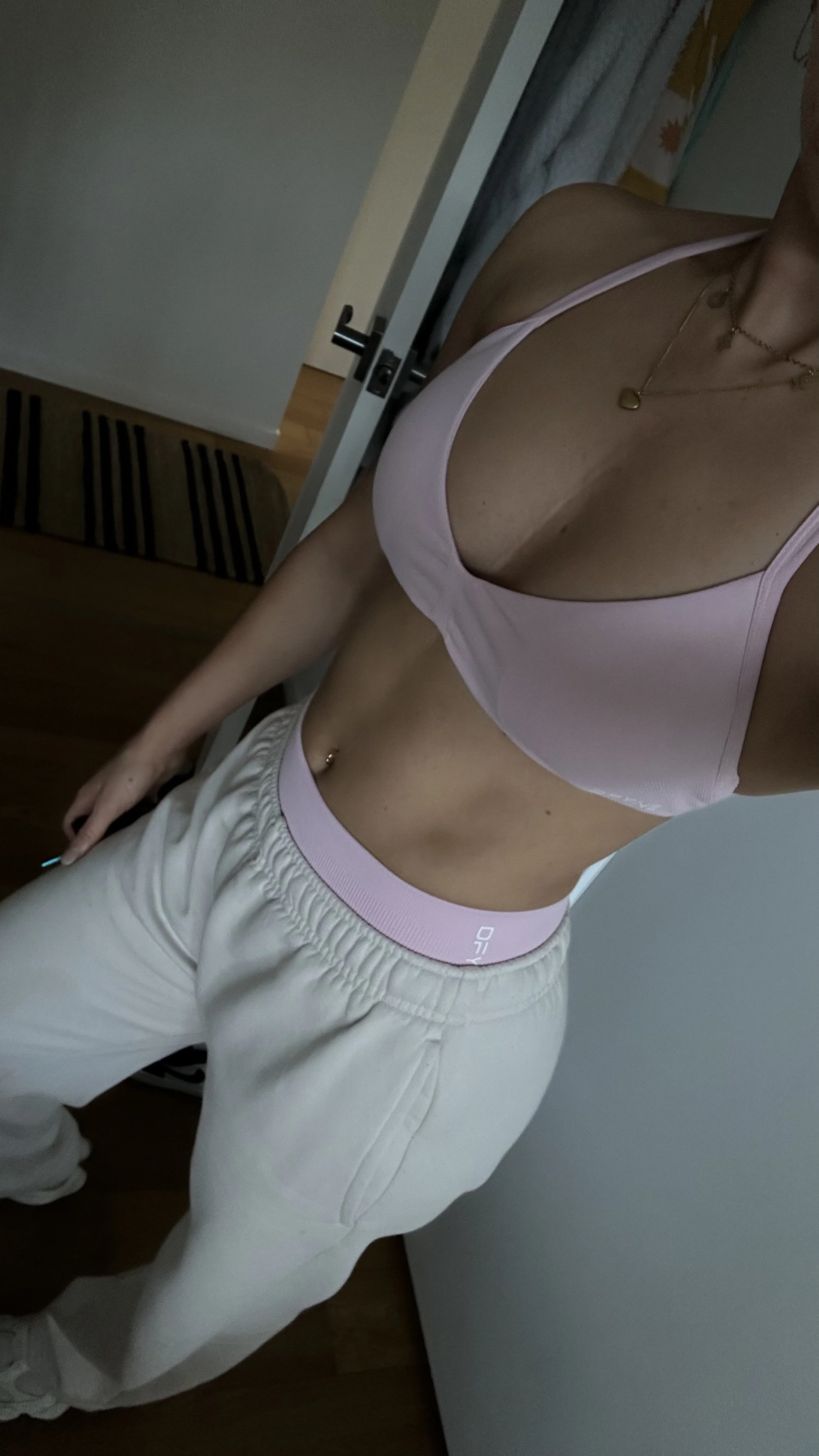 A woman wearing a pink sports bra and beige sweatpants, standing in front of a mirror, with a door and floor visible in the background.