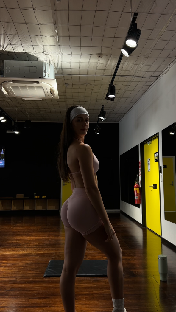 Woman in gym attire standing in a fitness studio, wearing a headband and pink shorts, with yellow doors and wooden floor visible.