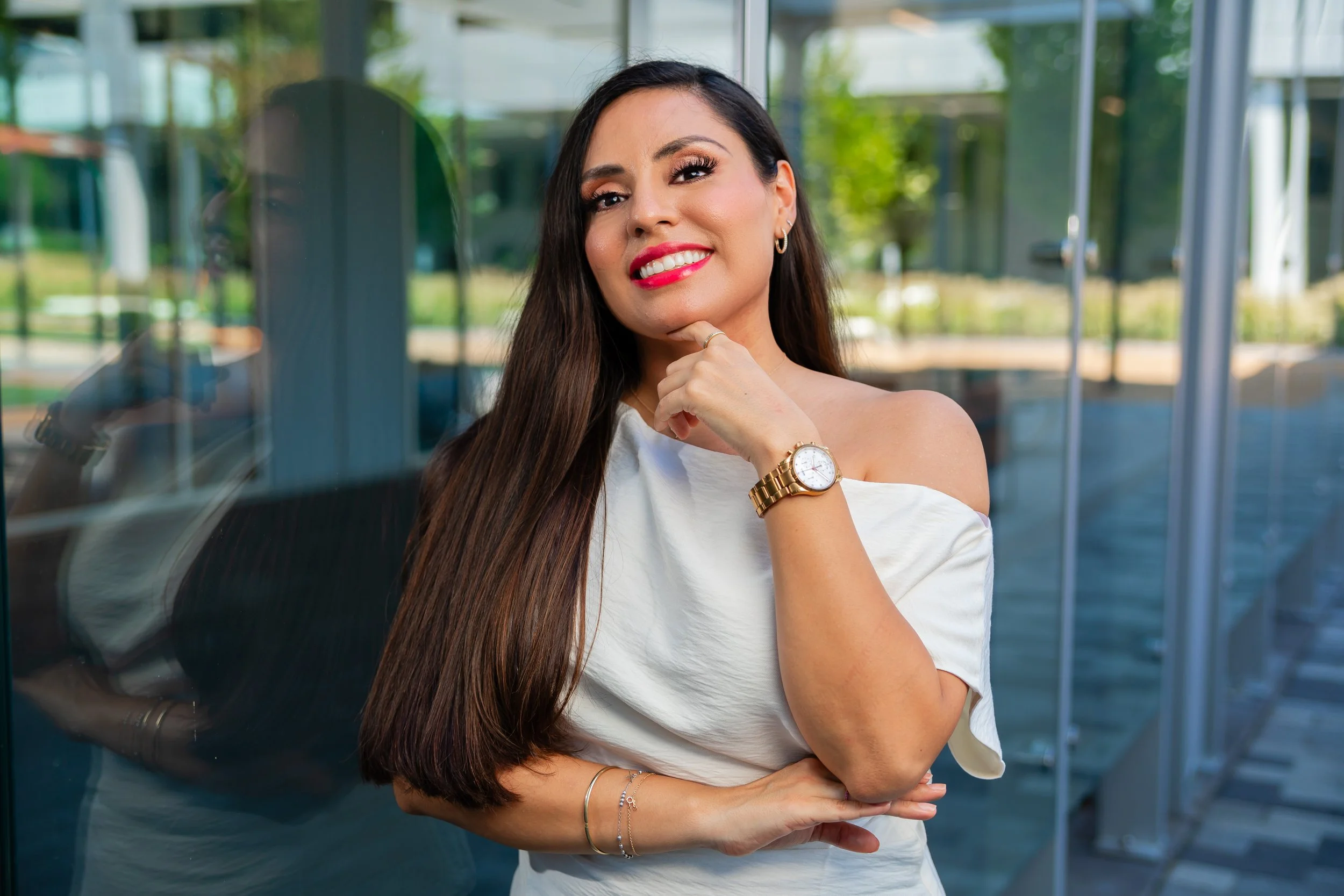 Woman with long brown hair, wearing a white off-shoulder top and gold watch, standing outside near a glass building, smiling and posing with her hand on her chin.