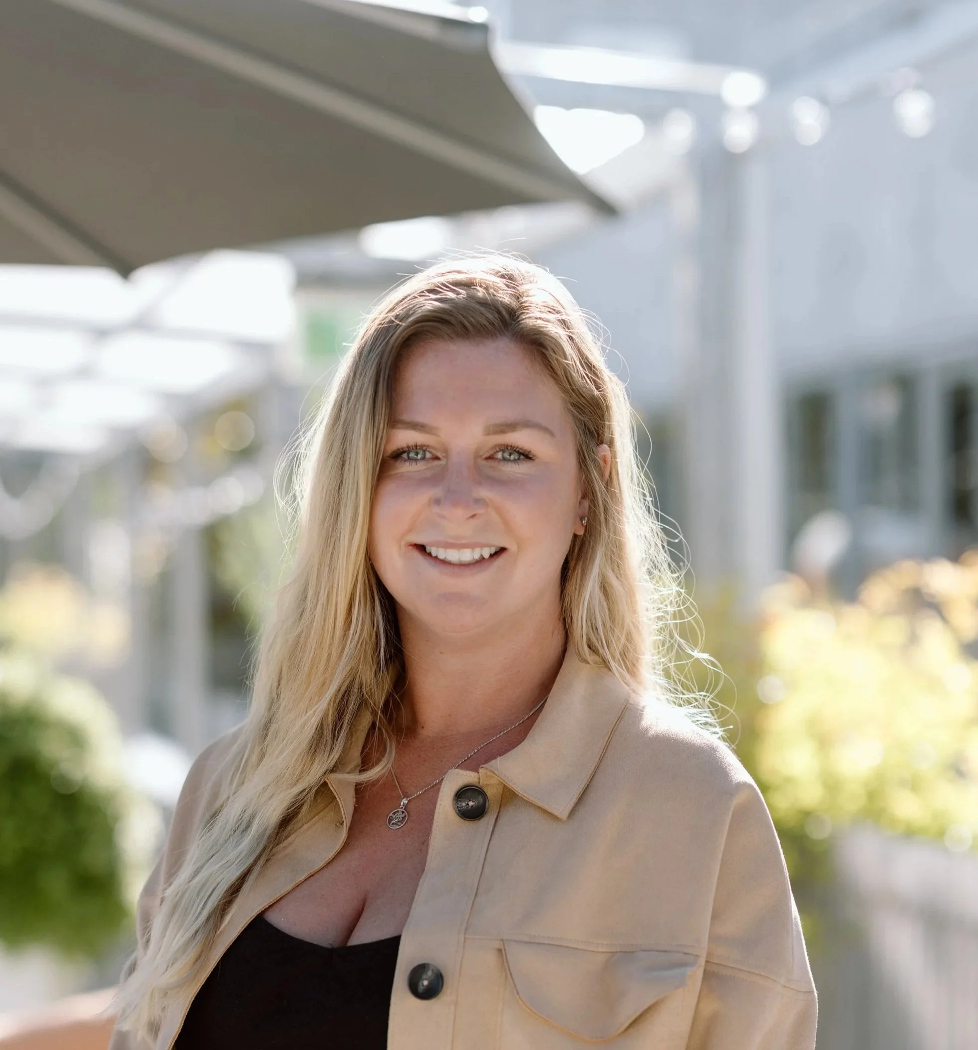 A smiling woman with long blonde hair, wearing a beige jacket and black top outdoors on a sunny day.