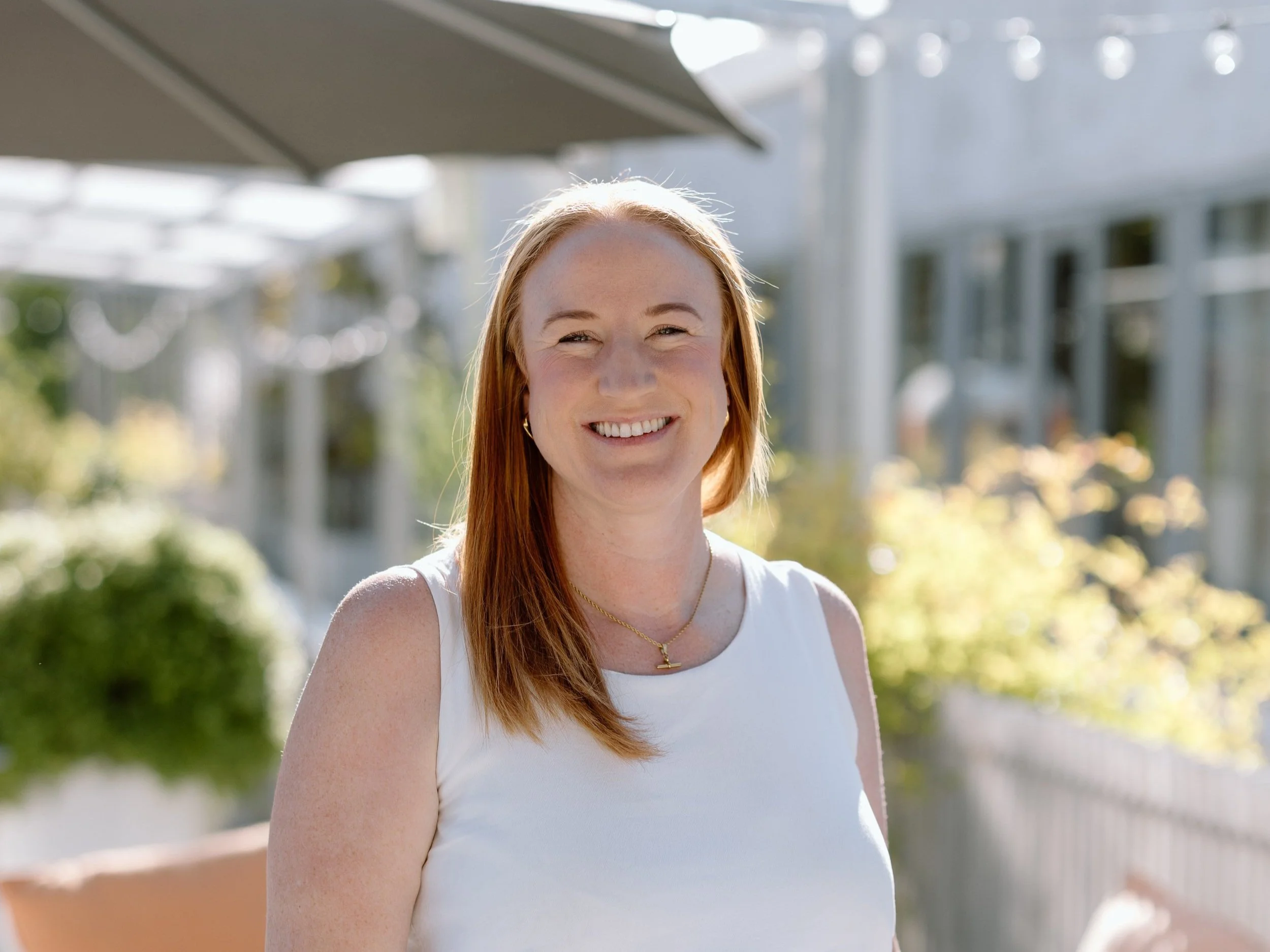 A smiling woman with long red hair wearing a white sleeveless top, standing outdoors in bright sunlight with an umbrella and a building in the background.