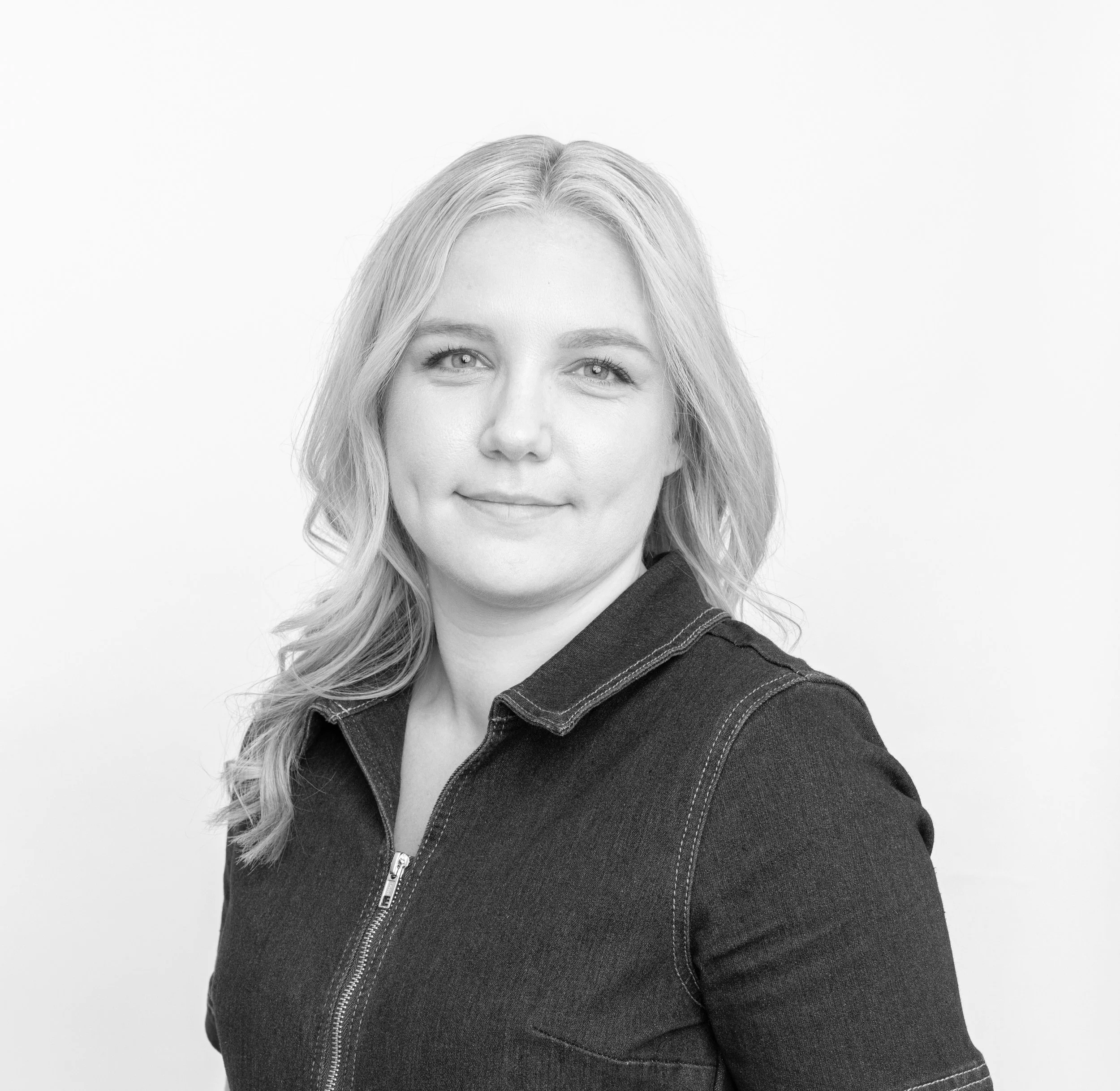 Black and white portrait of a young woman with wavy, shoulder-length hair, smiling slightly, wearing a dark zip-up denim jacket against a plain light background.